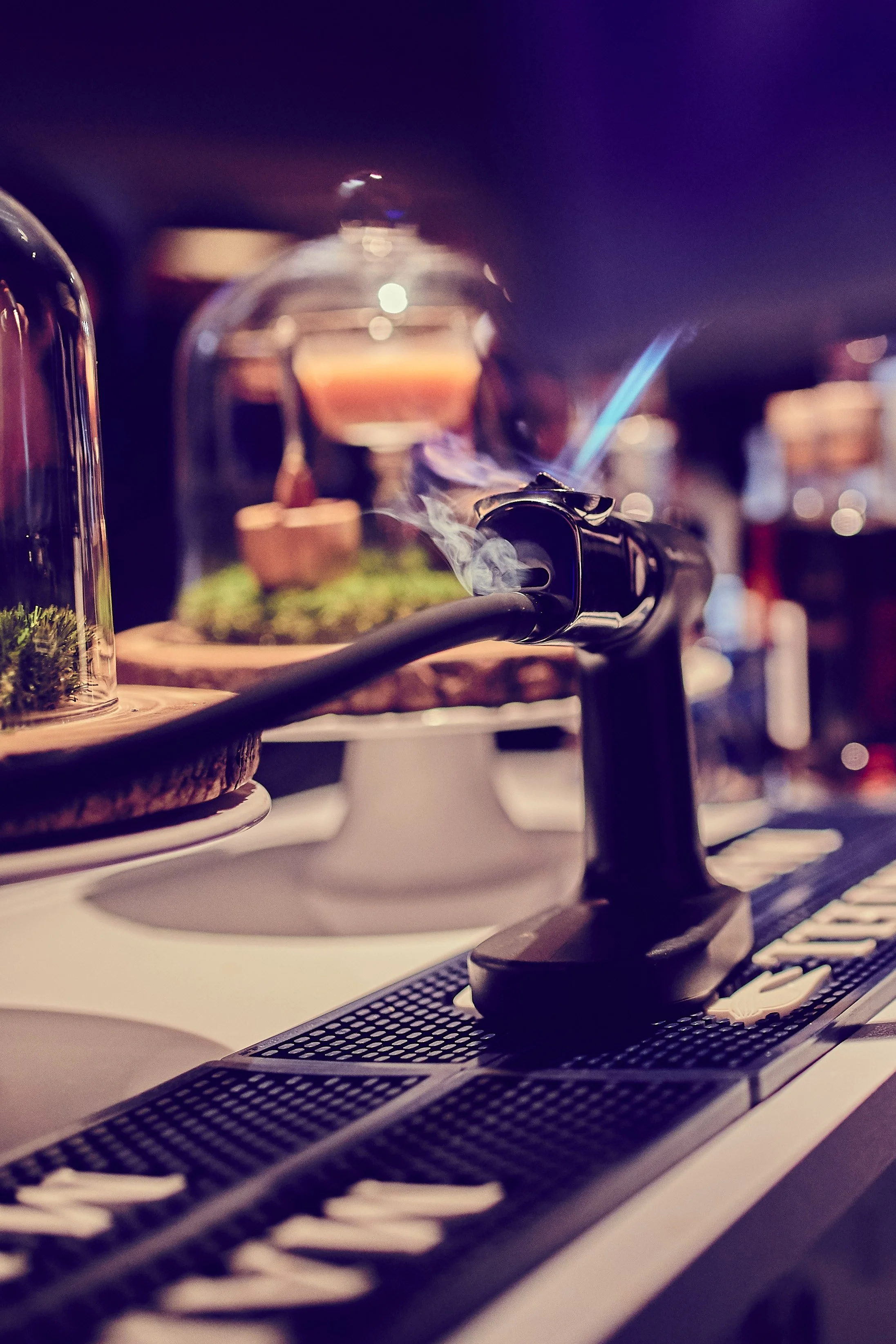 Close-up of an electric lighter with smoke, placed on a bar or countertop with blurred background of bottles and bar items.