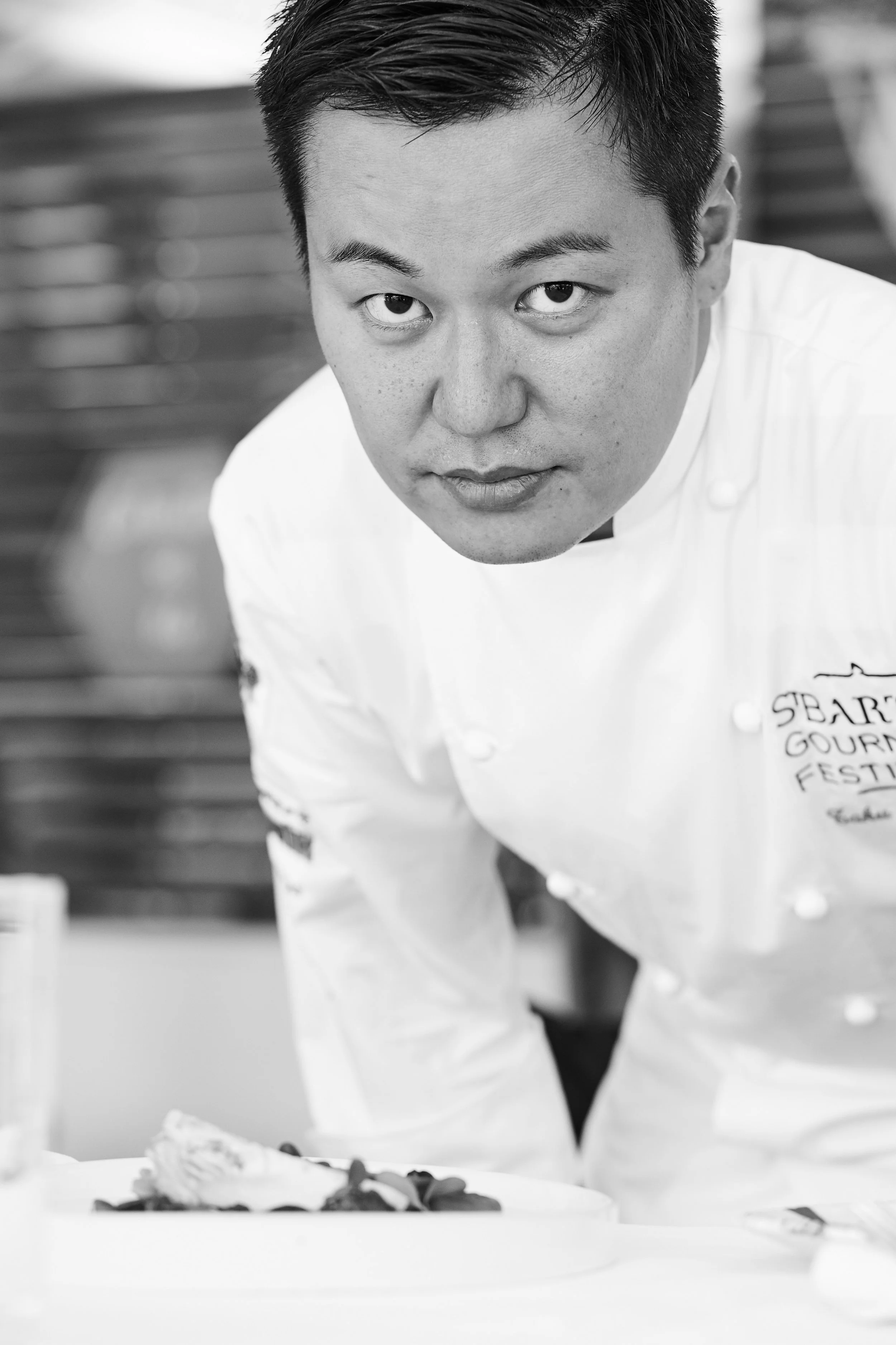 Black and white photo of a male chef looking directly at the camera, wearing a white chef's coat with some embroidered text on the chest, leaning over a table with a dish.