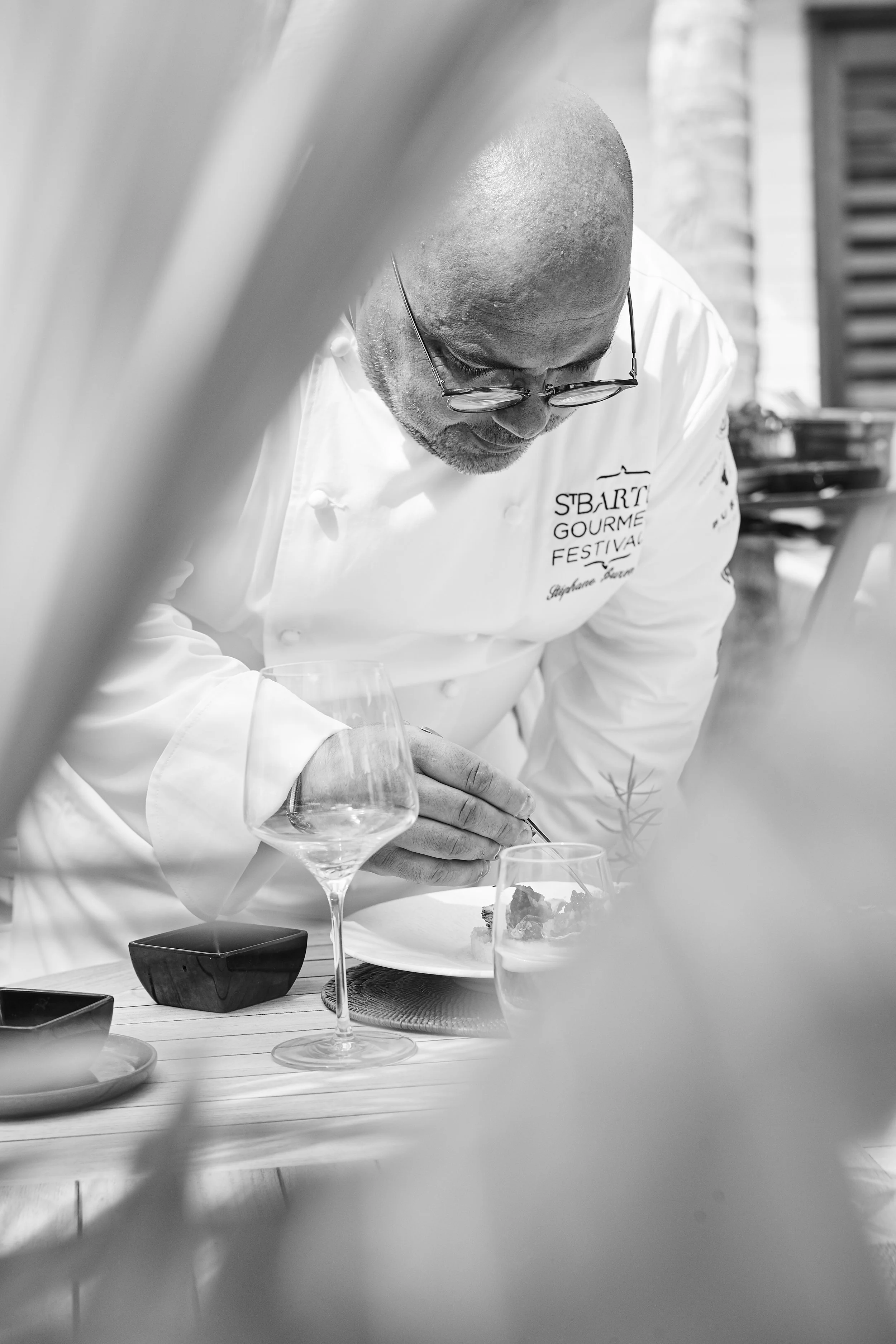A chef wearing glasses and a white uniform preparing a dish at a restaurant or culinary event, with wine glasses and tableware on the table.