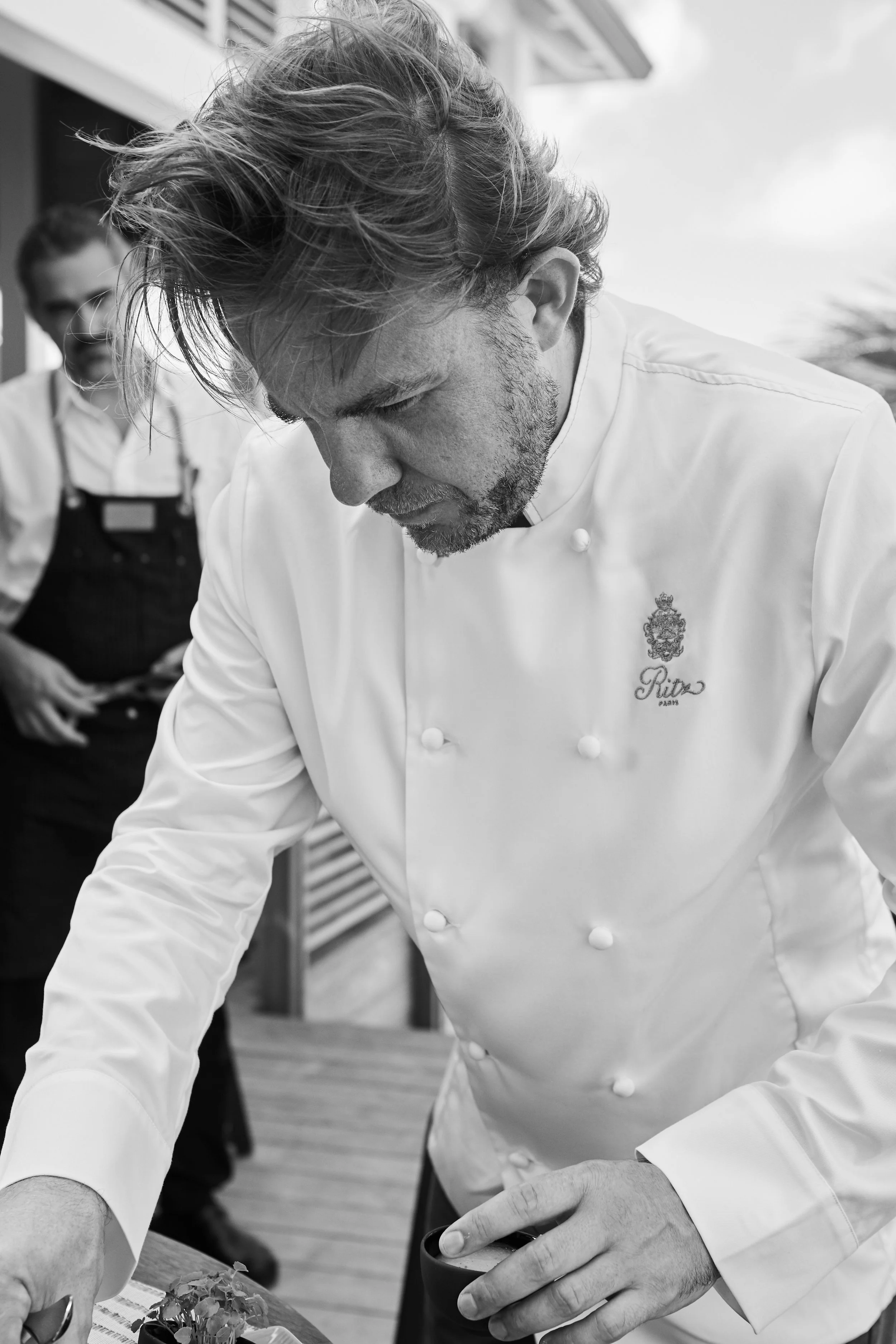 A male chef with disheveled hair and a beard, dressed in a white chef's coat, is arranging food on a table with a woman in black apron behind him, outdoors under a cloudy sky.