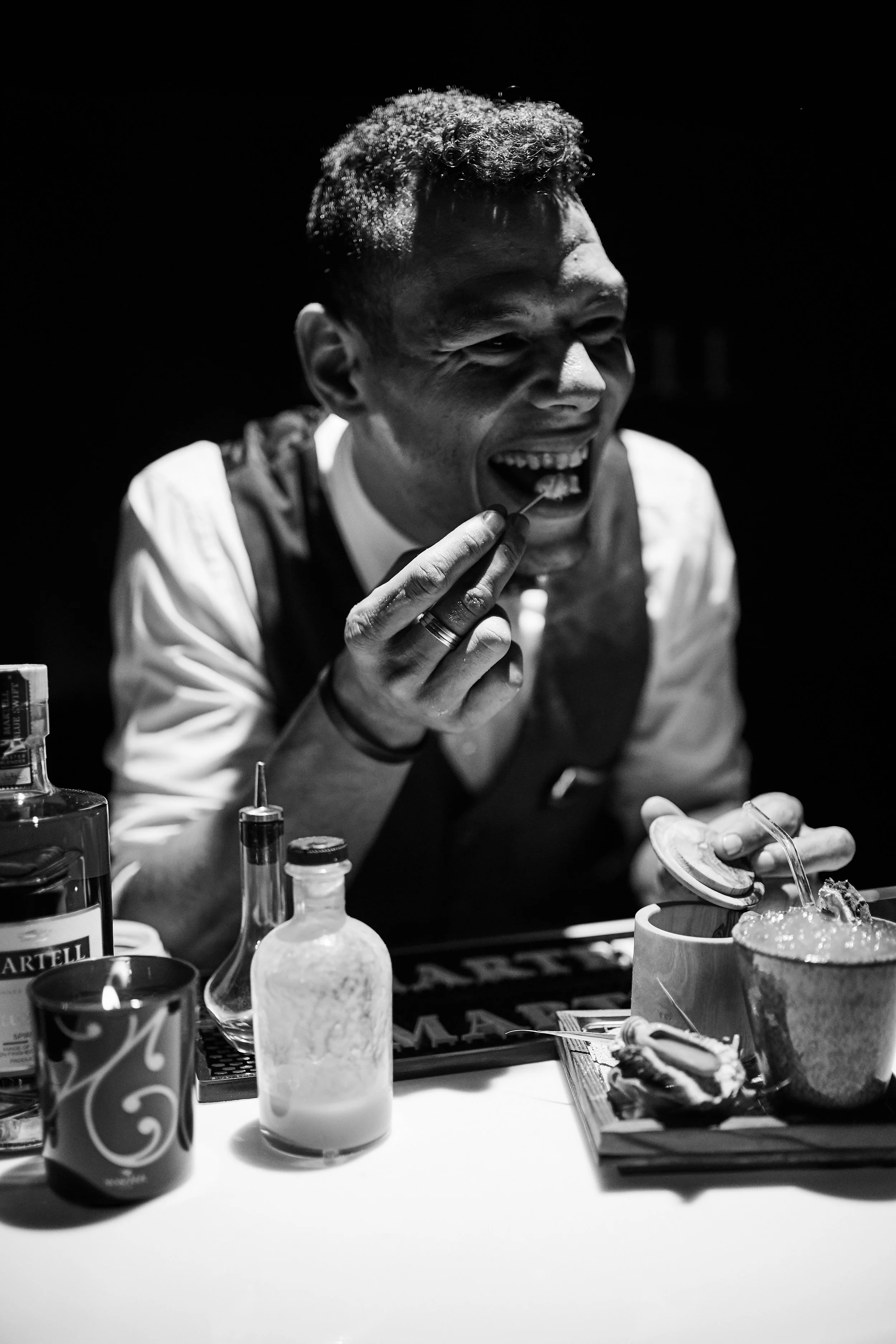 A man with short, curly hair in a vest and white shirt laughing while taking a bite of food. On the table are bottles, a mug, and bowls with food. The image is in black and white.