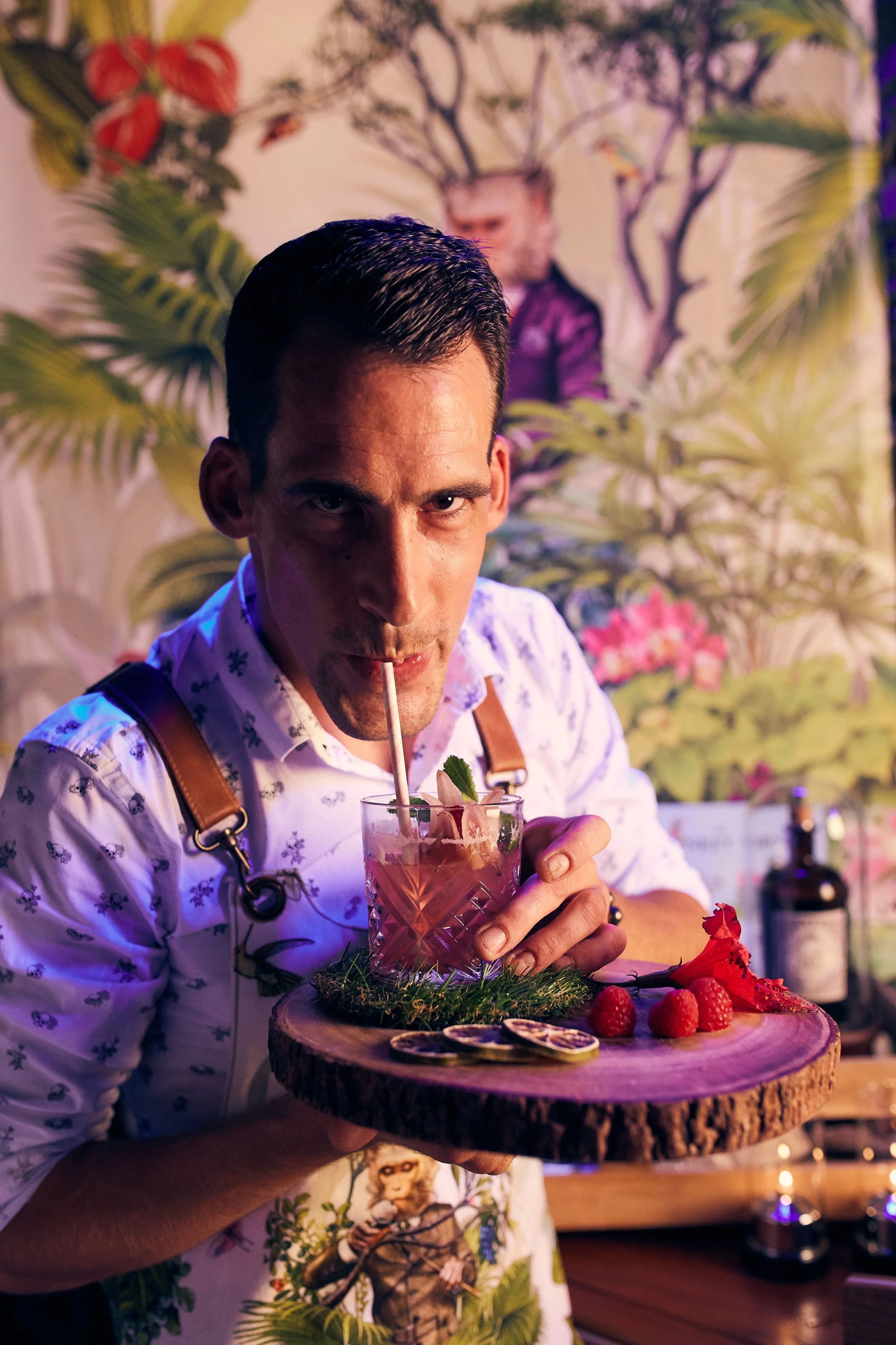 A man holding a wooden tray with a pink cocktail garnished with berries, lemon slices, and mint leaves. The background features lush plant decor and a mural of a woman with deer antlers.