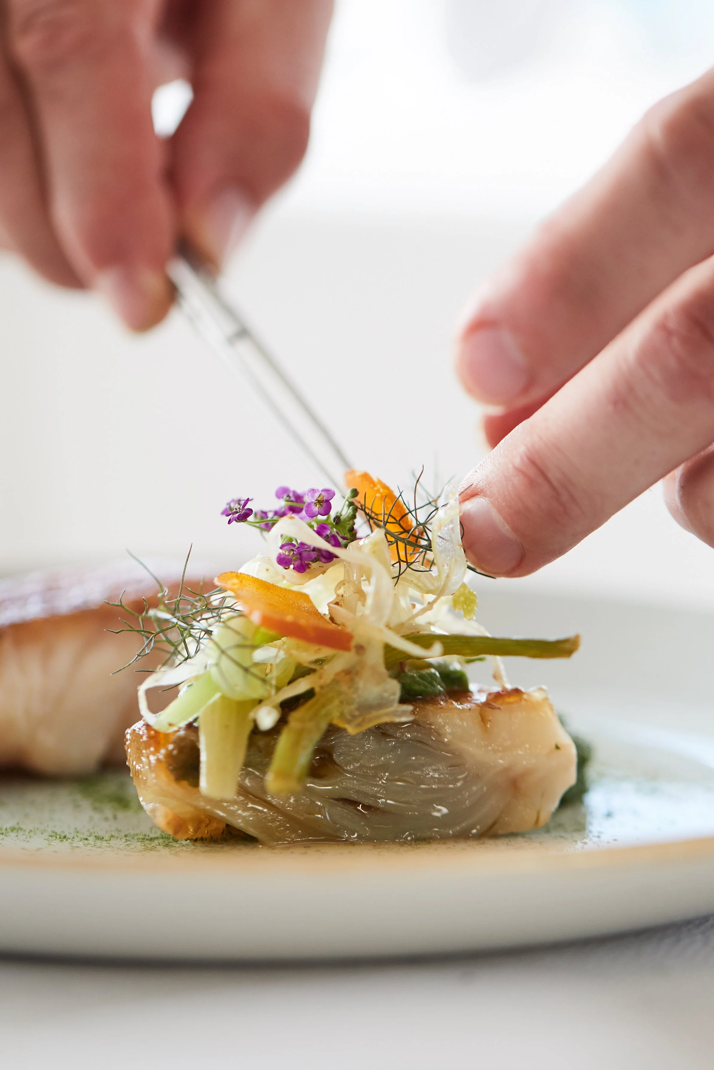 Chef garnishing a plated gourmet fish dish with edible flowers and microgreens.