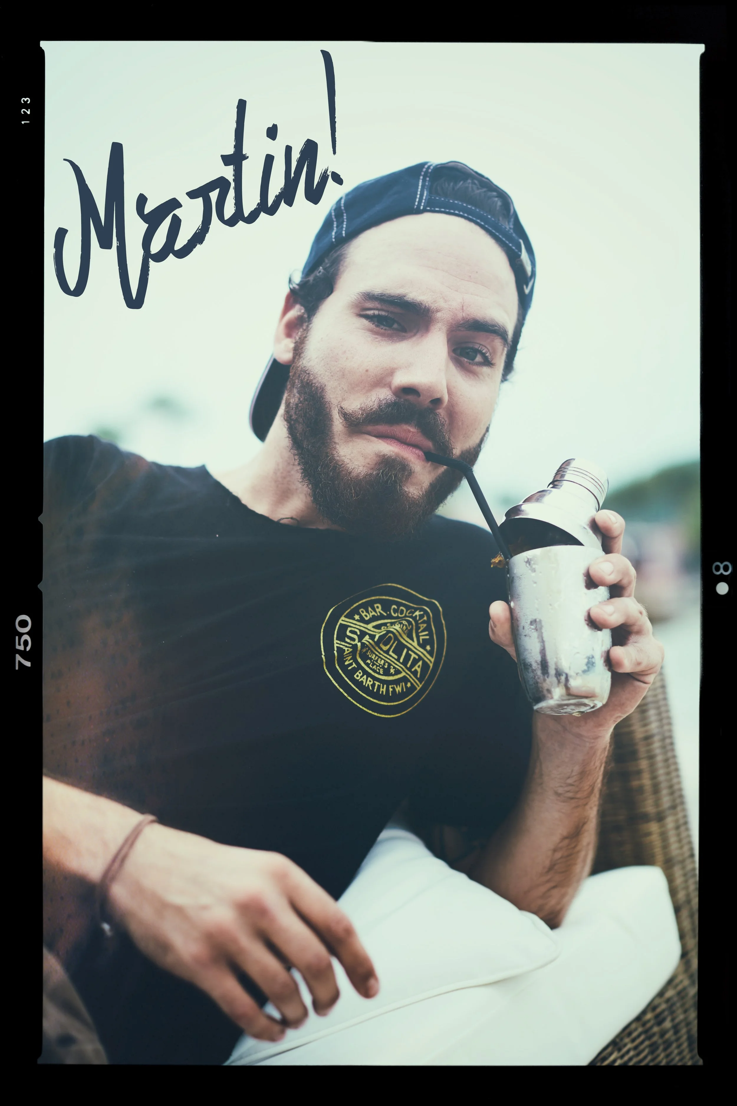 A man with a beard, wearing a black cap backwards and a black t-shirt, is holding a stainless steel flask and drinking from a straw. The shirt has a yellow logo that says 'Solita' and other text. The man is smiling slightly and looking at the camera.