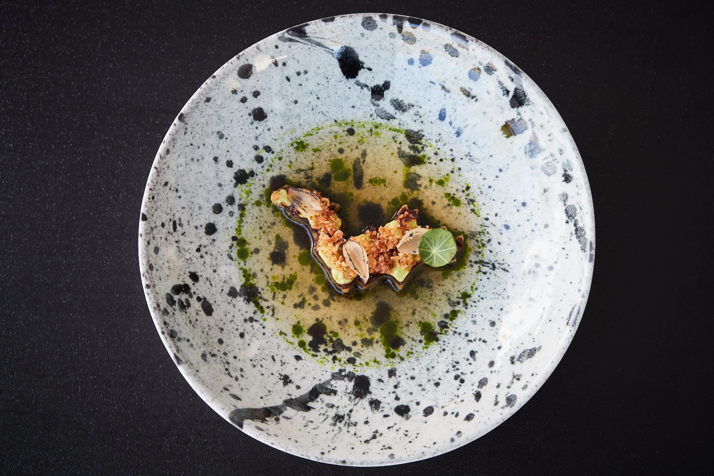 A modern, artistic plated dish with a small portion of food, garnished with a green leaf, on a speckled ceramic plate, set on a black surface.