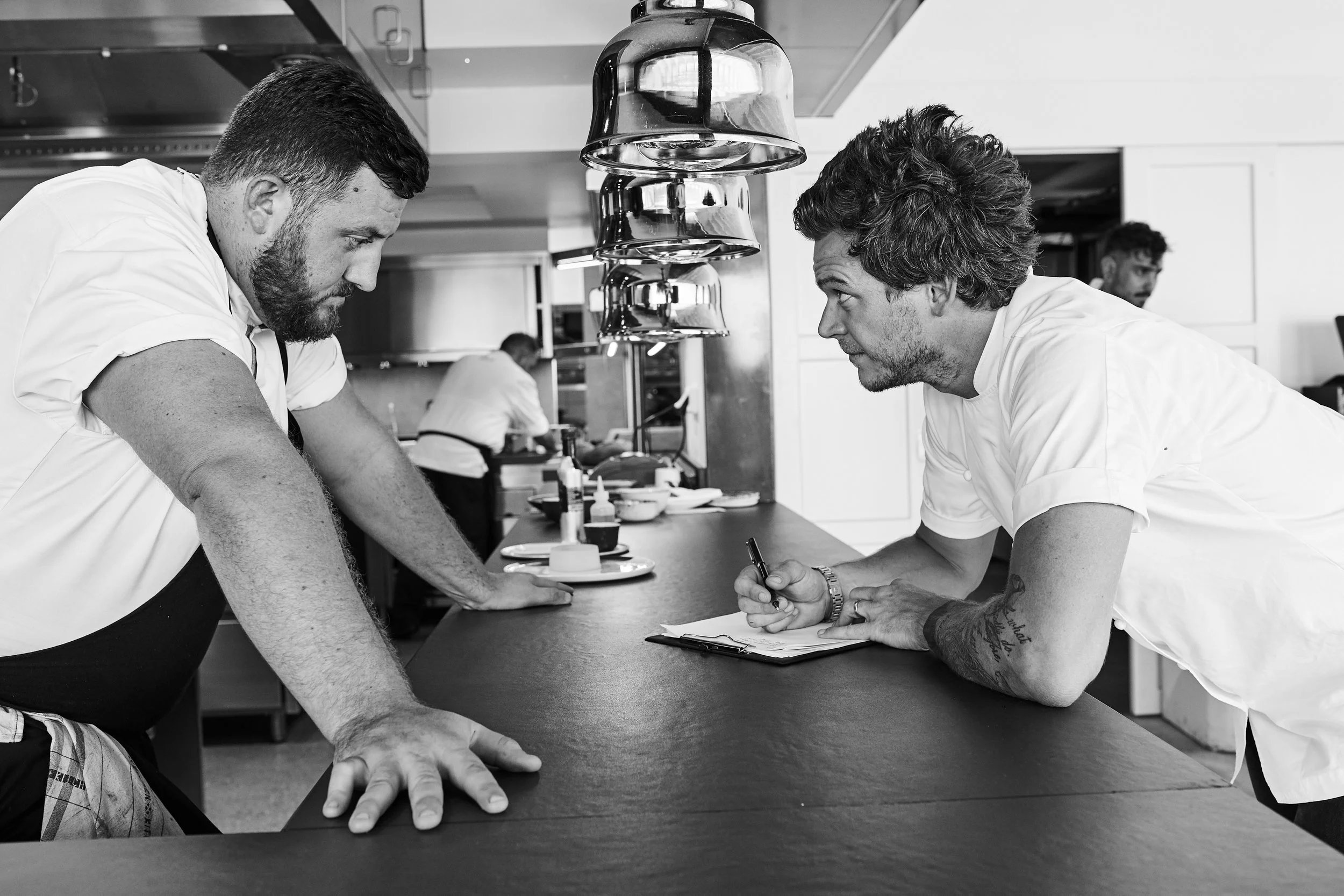 Two male chefs in white uniforms engaged in a discussion at a kitchen counter, with other kitchen staff working in the background.