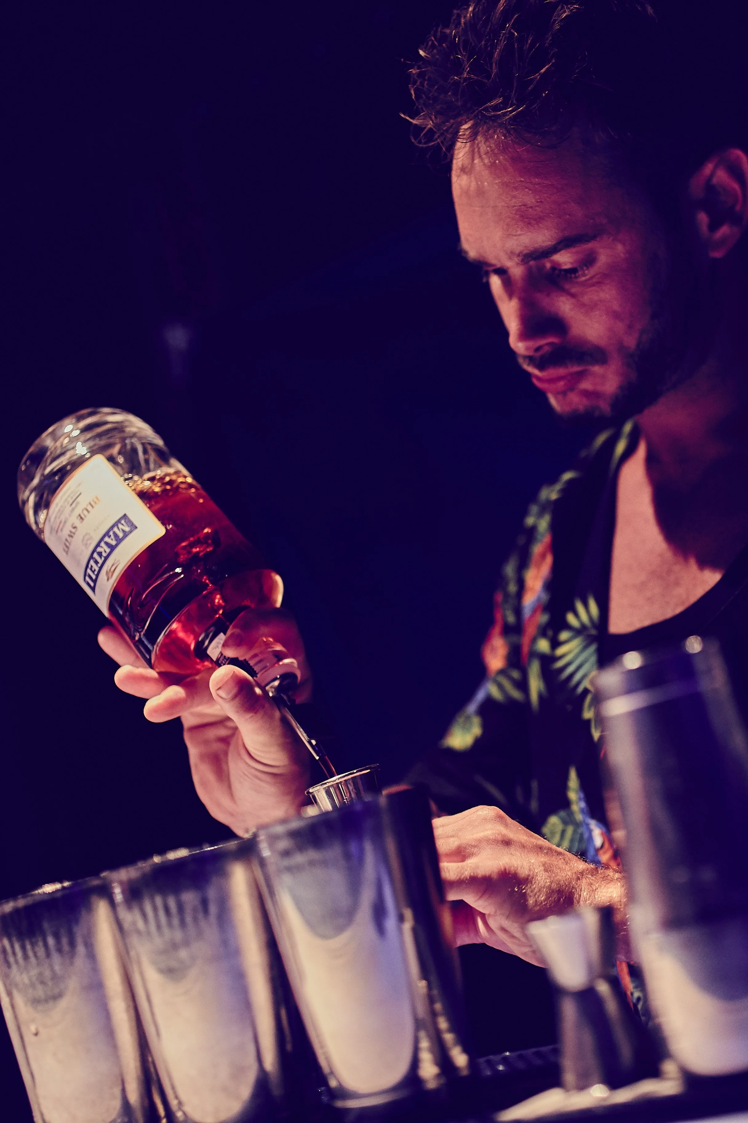 A man pouring a pink-colored liquor from a bottle labeled 'Marijuana' into a metal shaker, with a dark background.