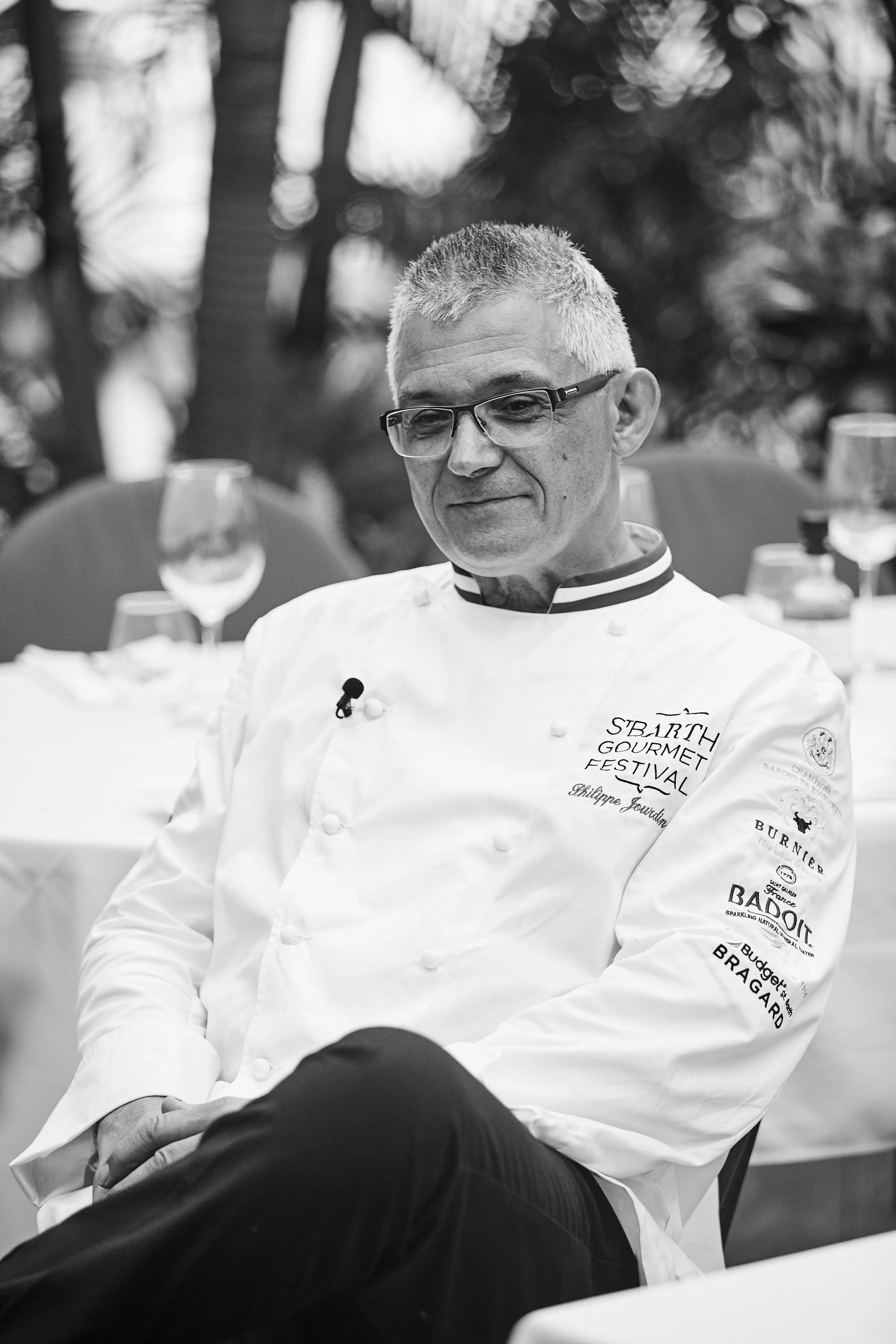 A male chef in a white chef's coat with logos and text, sitting at a dining table with glasses and cutlery in an outdoor setting.