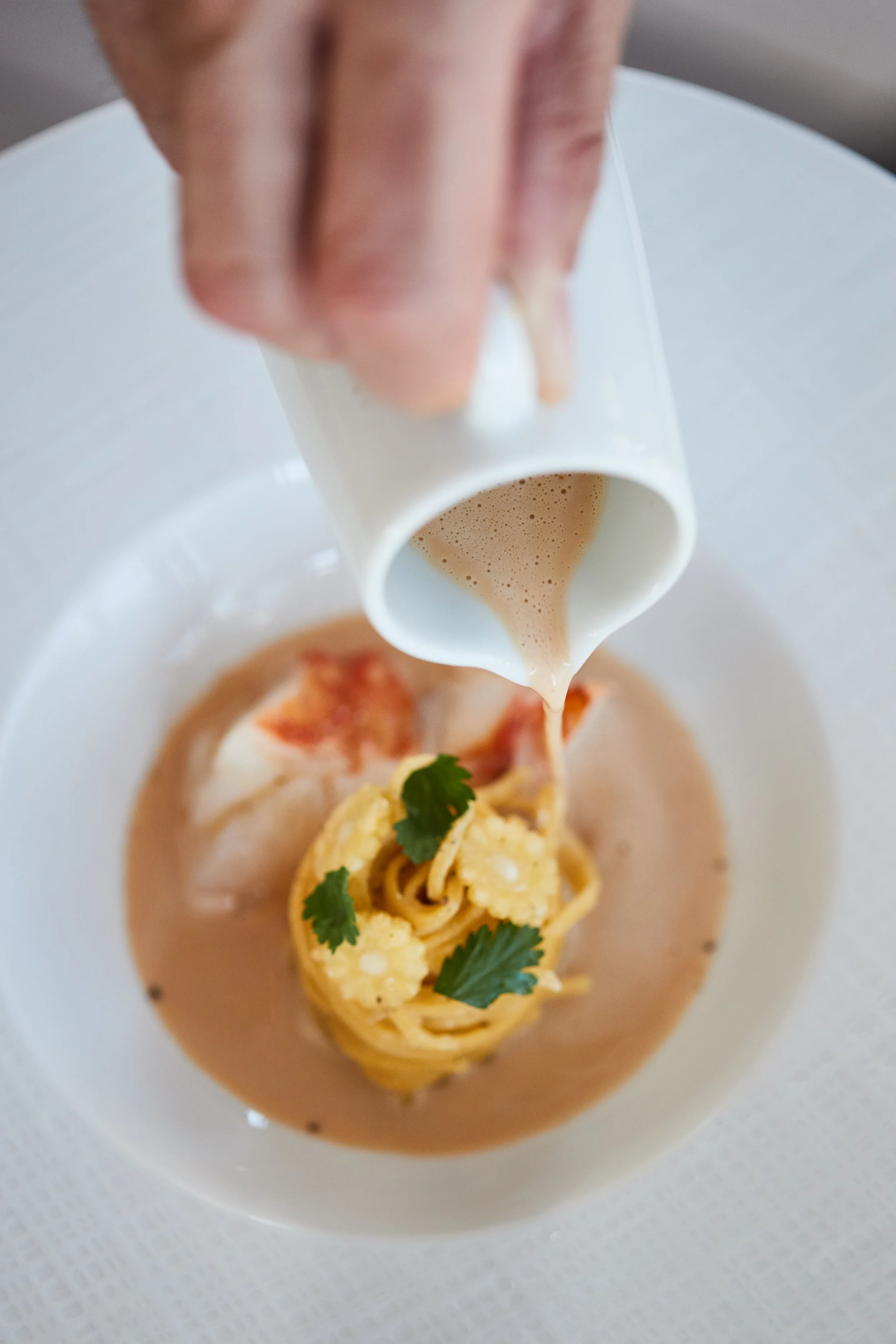 Pouring creamy soup over a plated pasta dish garnished with cilantro leaves.