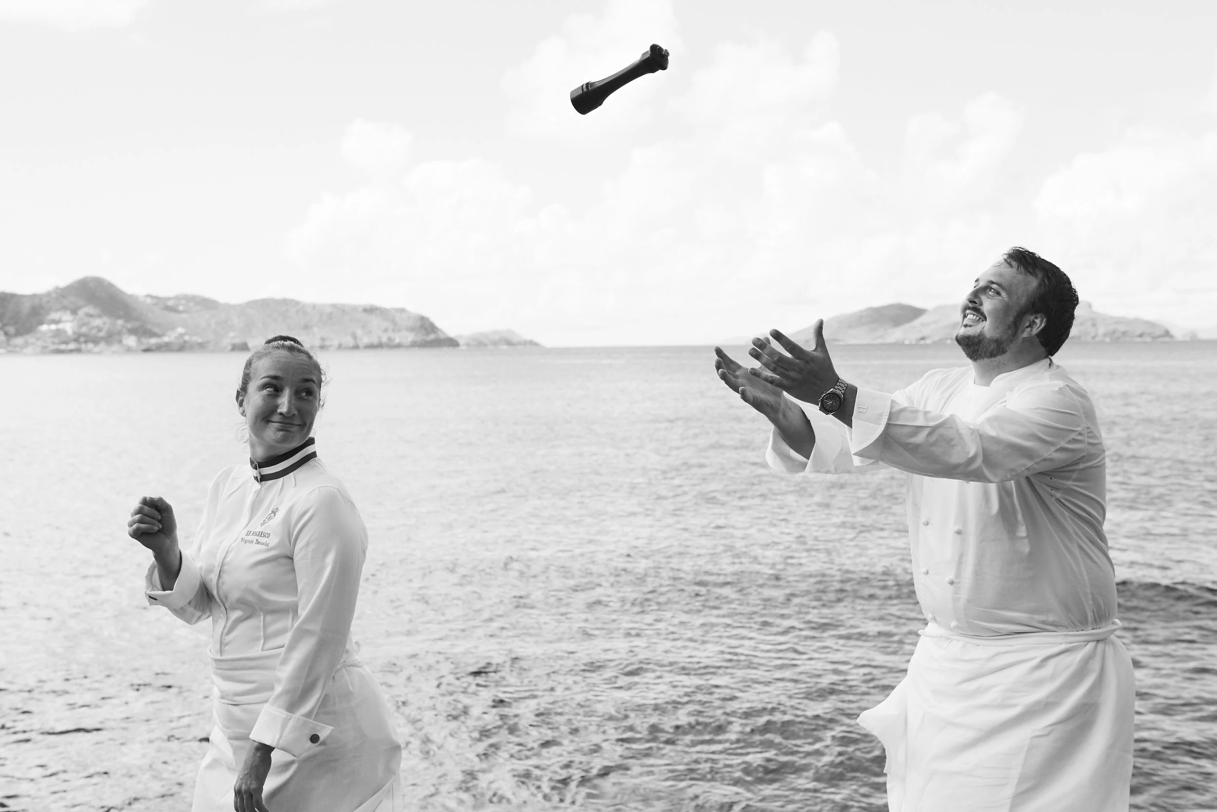 Two chefs in white uniforms outdoors near a body of water, one with a woman looking at him and the other tossing a bottle into the air.