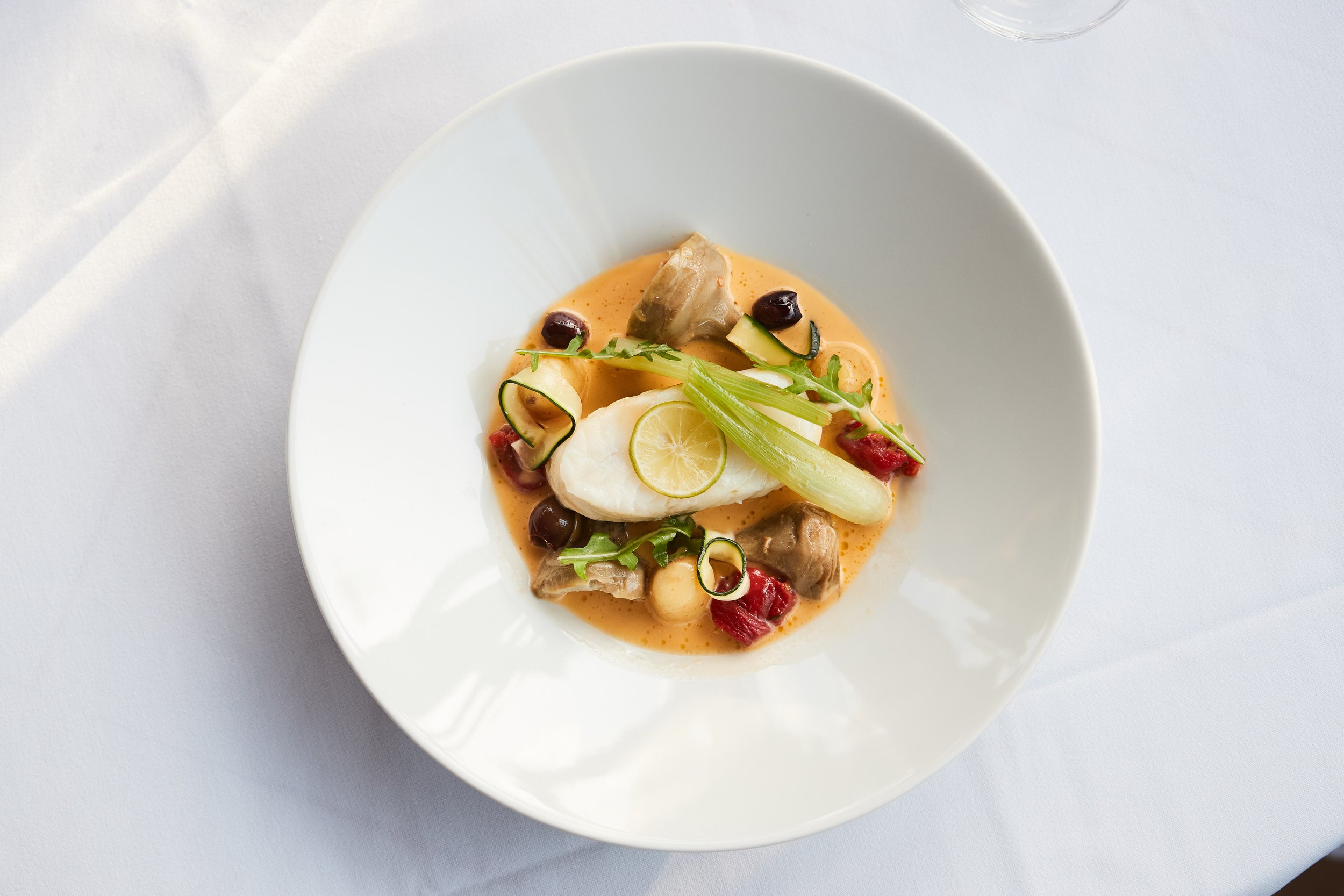 A plated gourmet dish featuring a poached egg, vegetables, and a creamy sauce, garnished with lemon slices and microgreens.