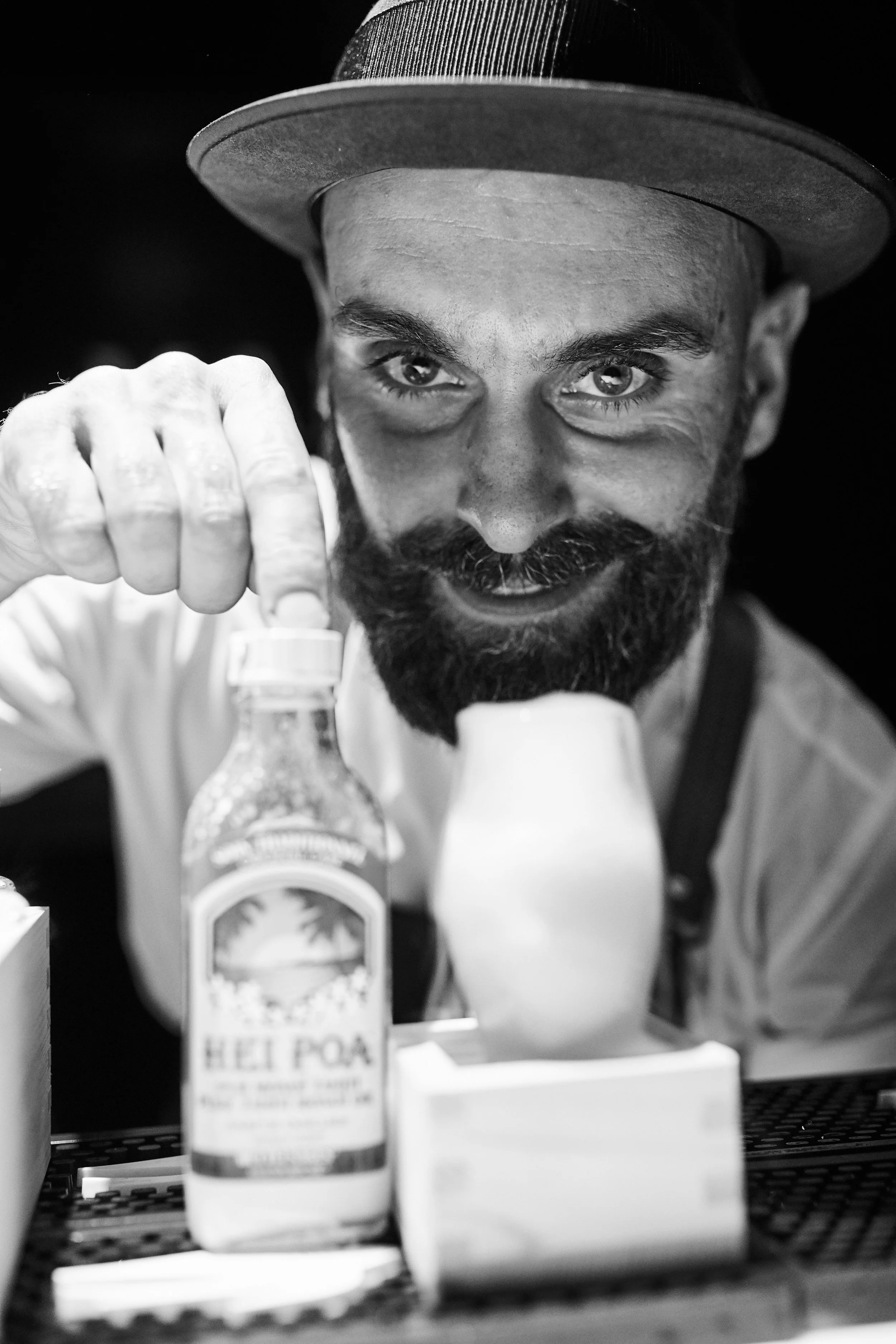 A man with a beard and a hat smiling at the camera, reaching toward a bottle of bottled water labeled "Help Pour," inside a cafe or bar setting.