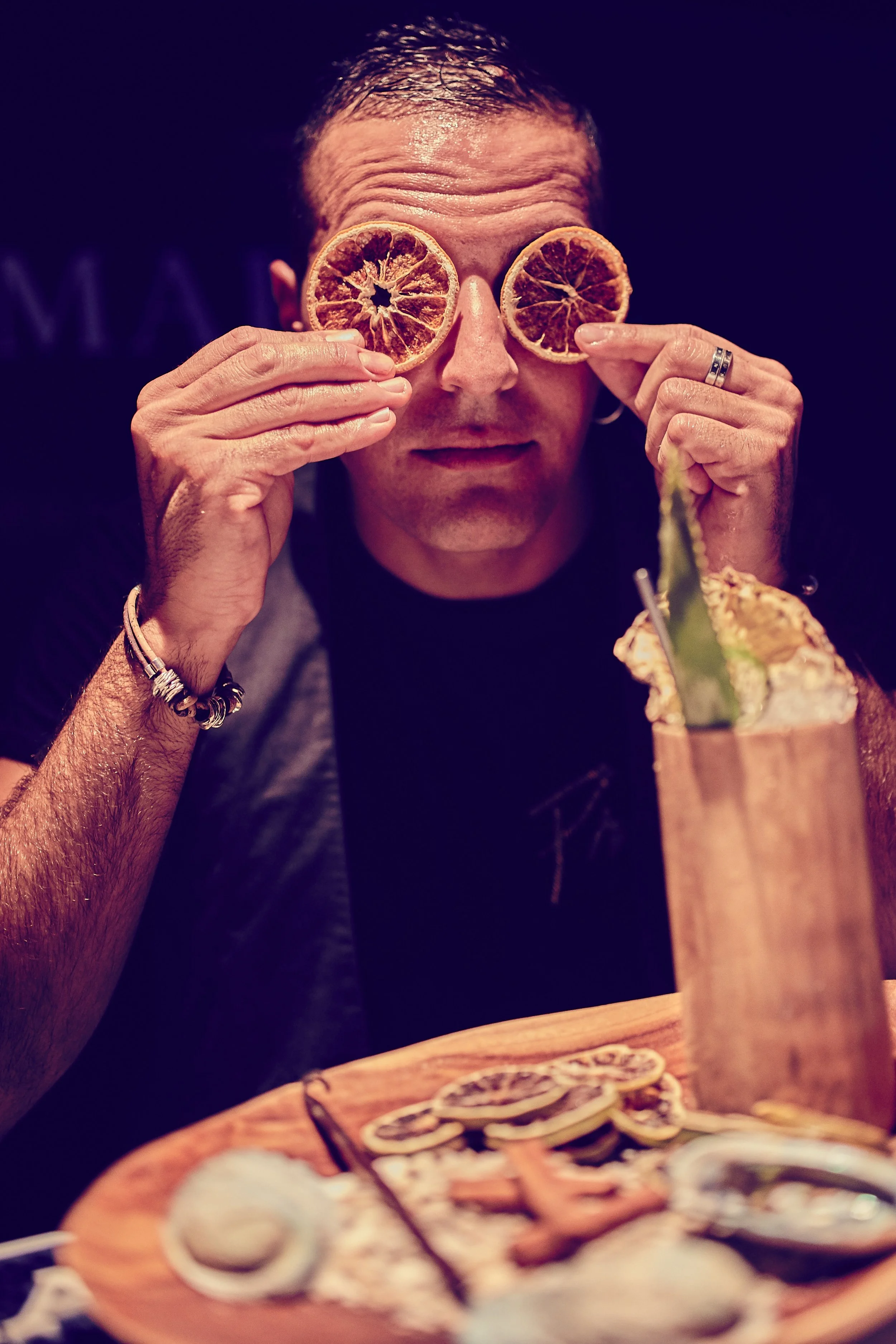 A man holding dried orange slices in front of his eyes at a restaurant or bar. There is a cocktail with a slice of aloe vera on a wooden table in front of him, along with a pizza and other dishes.