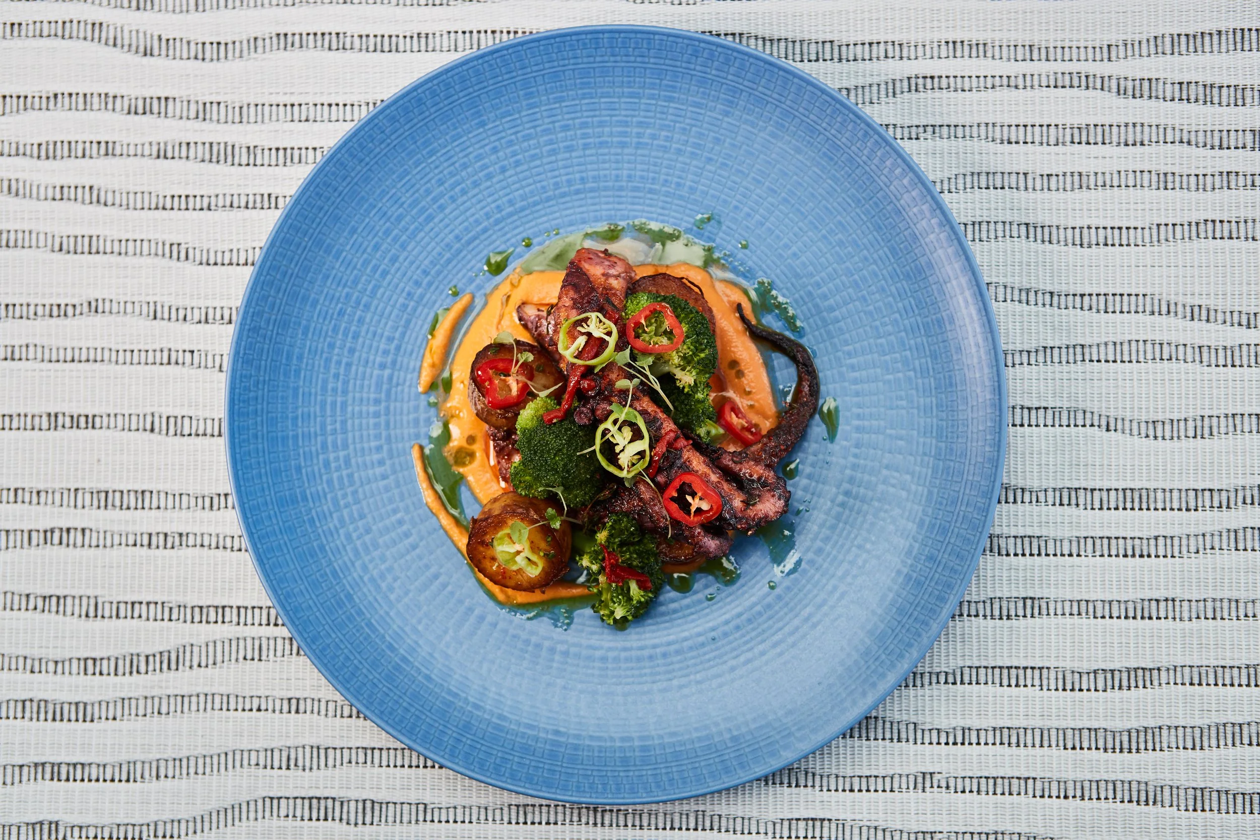A gourmet dish on a blue plate featuring grilled octopus, broccoli, cherry tomatoes, and sliced peppers, garnished with microgreens.