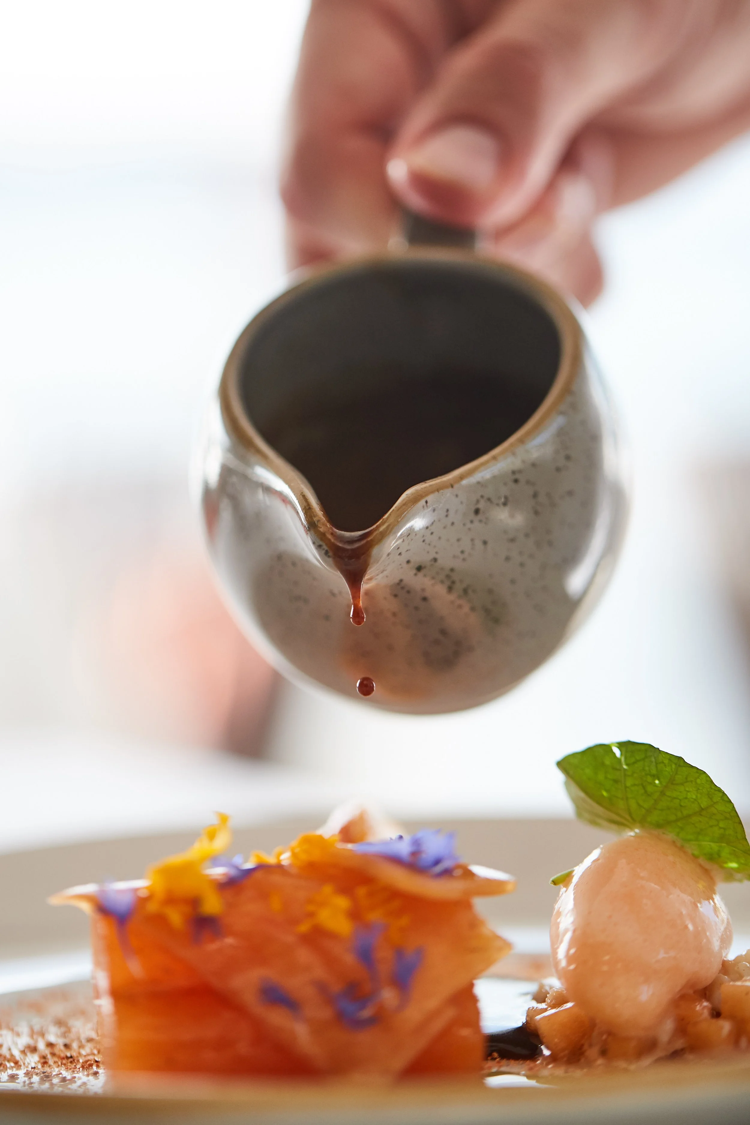 A hand pouring brown sauce from a pitcher onto a dessert plate that has a scoop of pink sorbet garnished with a green leaf, and a layered dessert topped with edible flowers and yellow and violet garnishes.