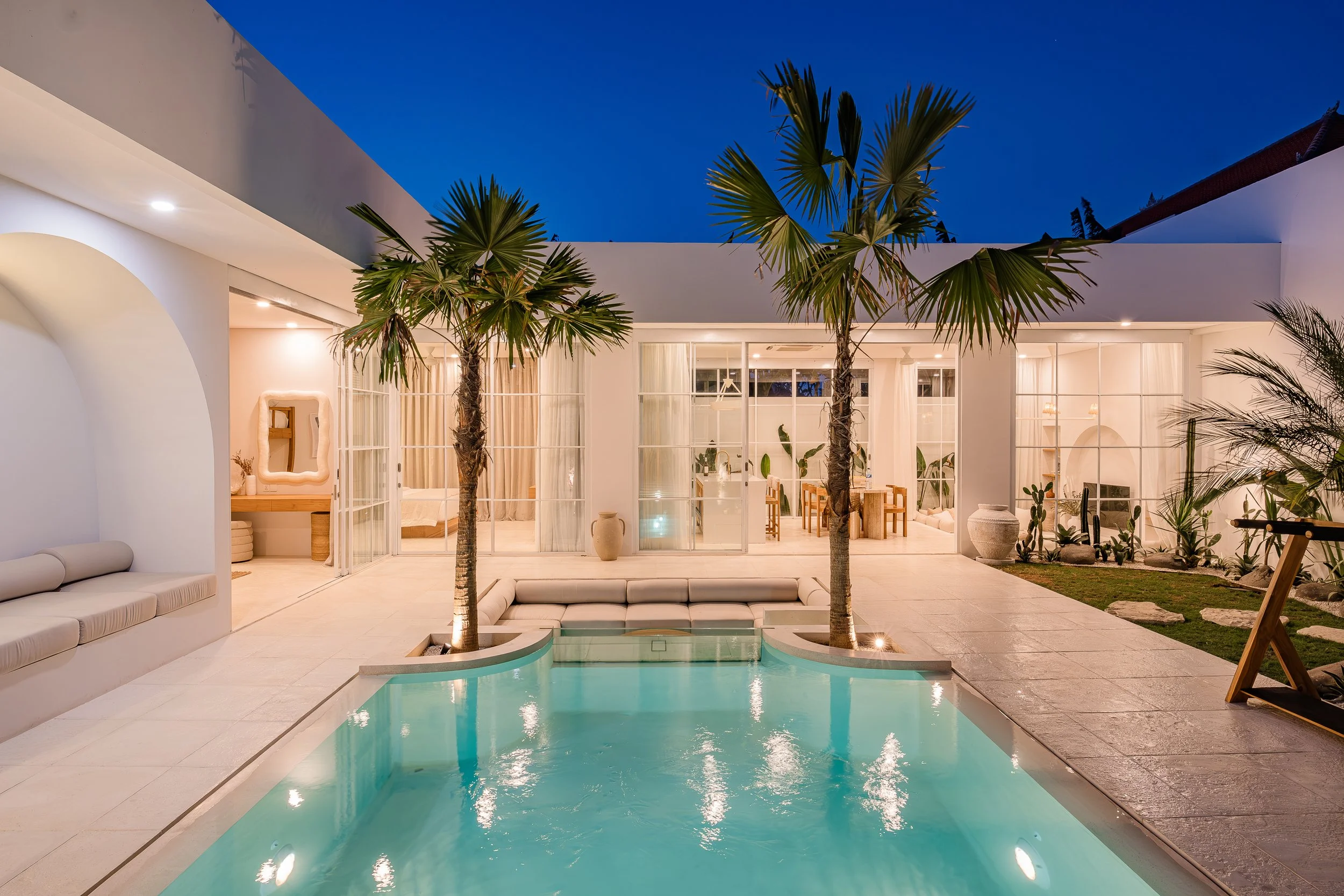 A modern house with a lit swimming pool in the front yard at night, featuring palm trees, outdoor seating, and interior views through large glass windows.