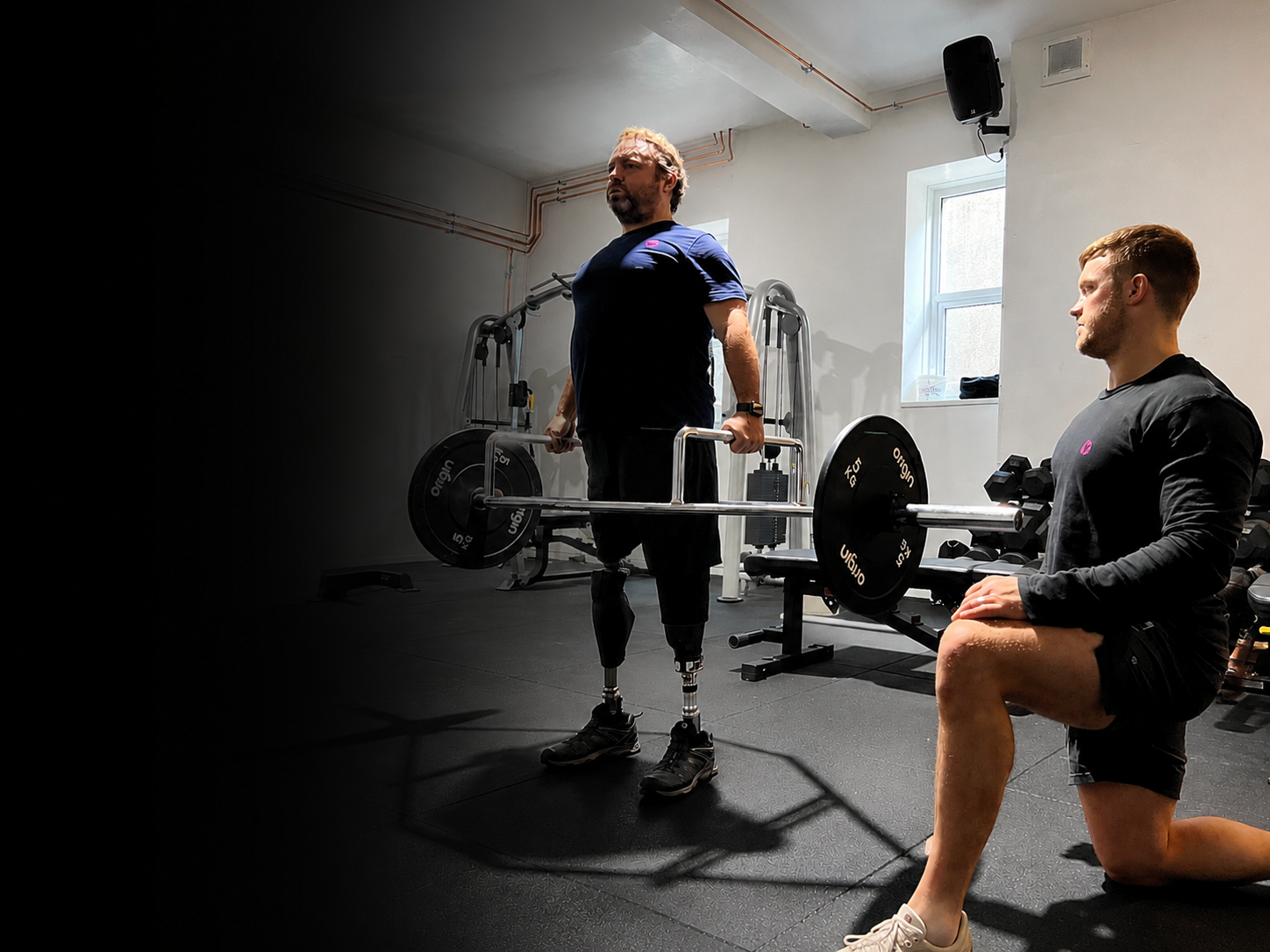 Personal trainer coaching an adaptive athlete during a strength training session at Pantheon Plymouth gym in Plymouth, Devon. The athlete performs a trap bar deadlift with bilateral prosthetic legs, while receiving inclusive one-to-one coaching.