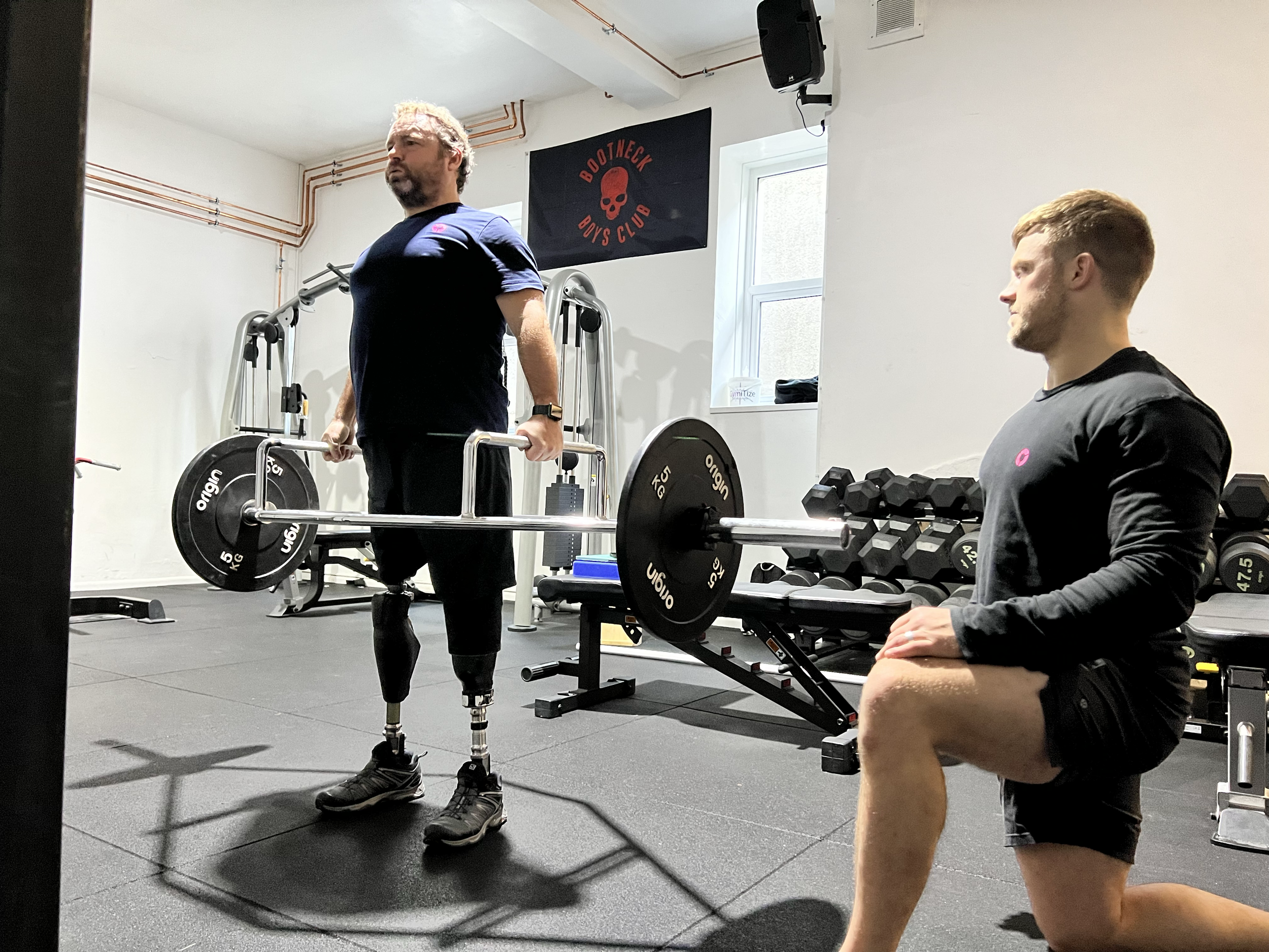 A man with prosthetic legs lifting weights at the gym while another man with short hair and a beard sits nearby. There is a banner on the wall with the text 'BootNeck Boys Club' and a skull logo. Gym equipment, including weights and a fitness machine