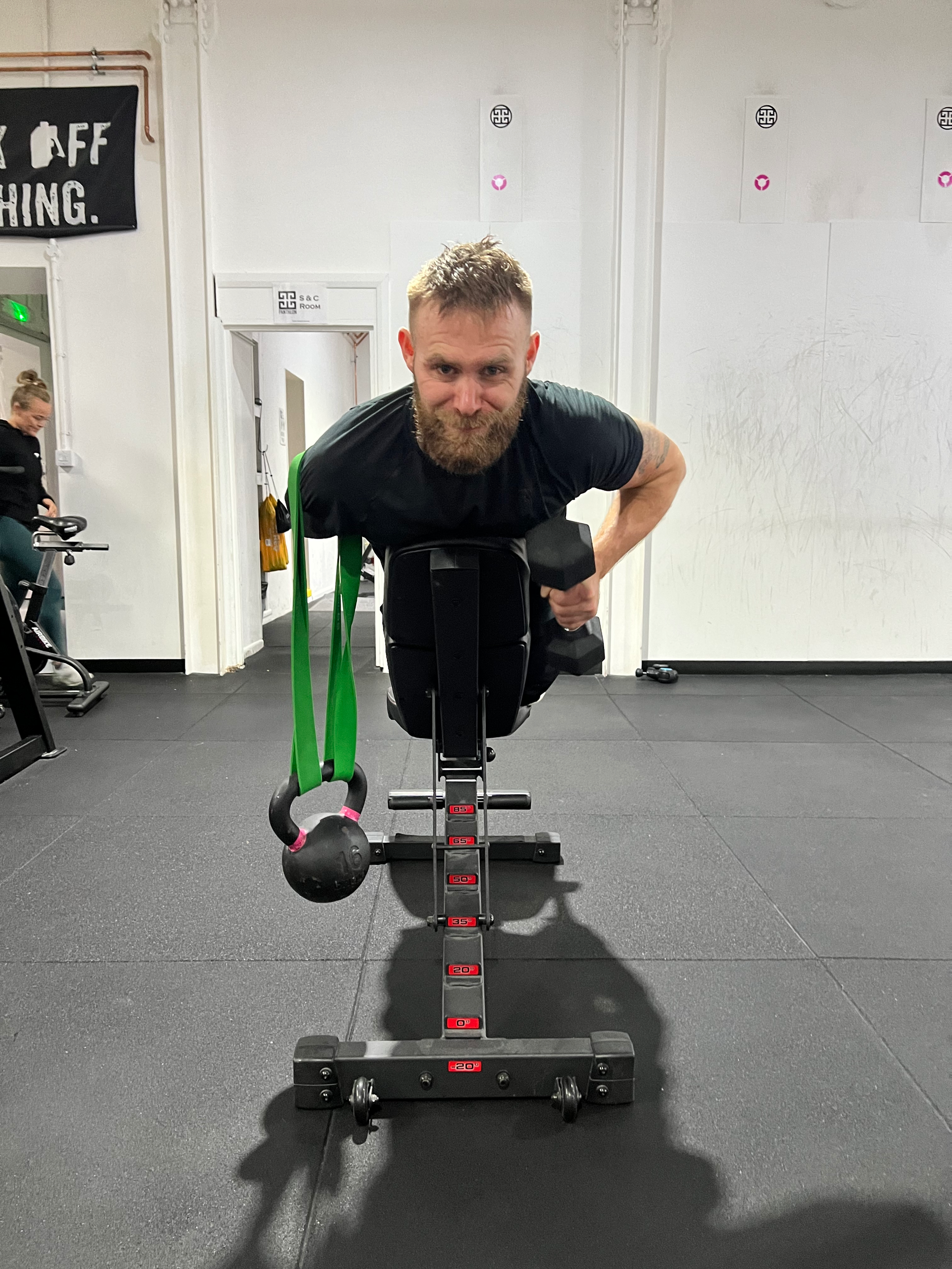 A man with a beard in a black shirt is performing a plank exercise on a layered platform in a gym. A kettlebell with pink handles is hanging from his waist, and his hands are on the platform, with a focused expression. The background shows another pe