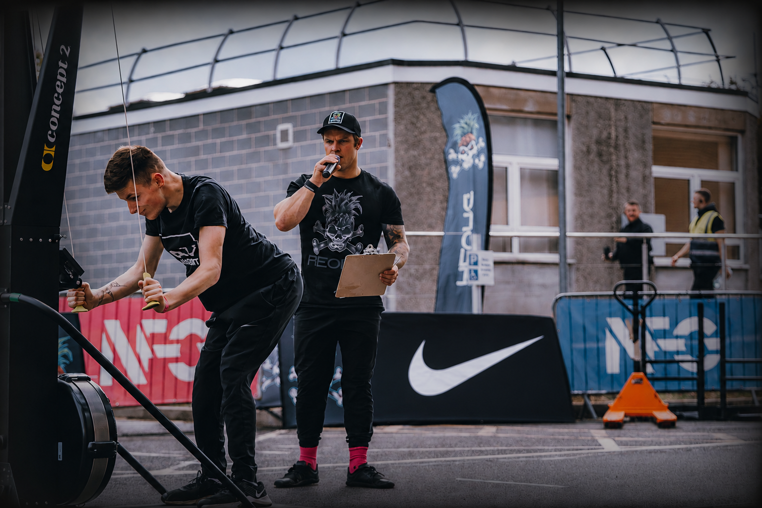Athlete competing on a Concept2 SkiErg machine during an outdoor adaptive fitness event while a coach provides guidance, with branded event signage and barriers in the background.
