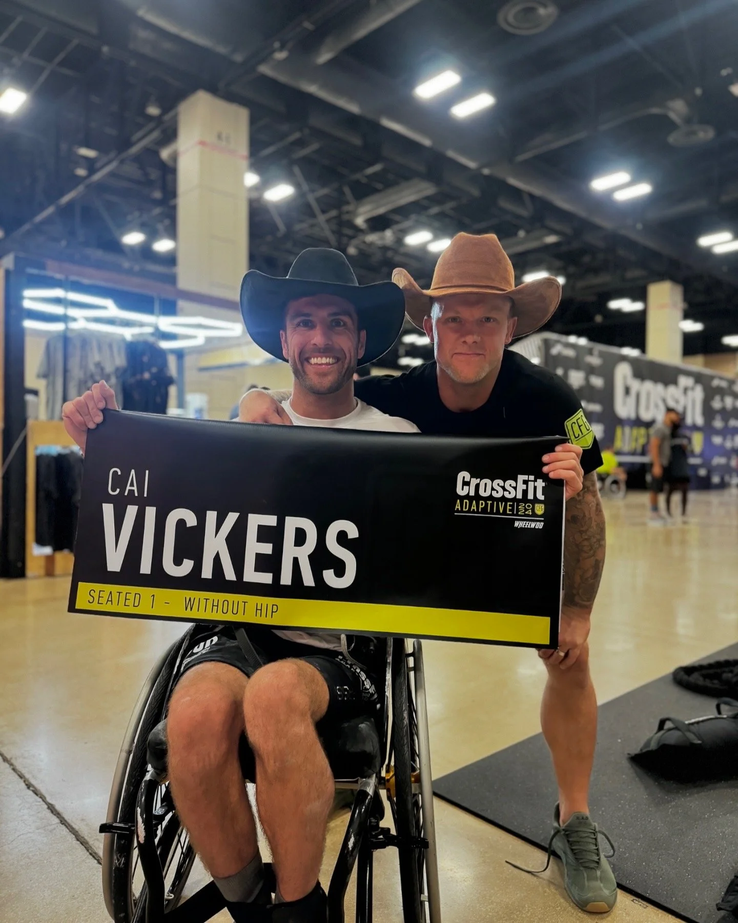 Two men wearing cowboy hats are indoors at a CrossFit fitness event. One man, sitting in a wheelchair, is holding a sign that reads "VICKERS SEATED 1 - WITHOUT HIP," and is smiling at the camera. The other man, standing beside him, has his arm around