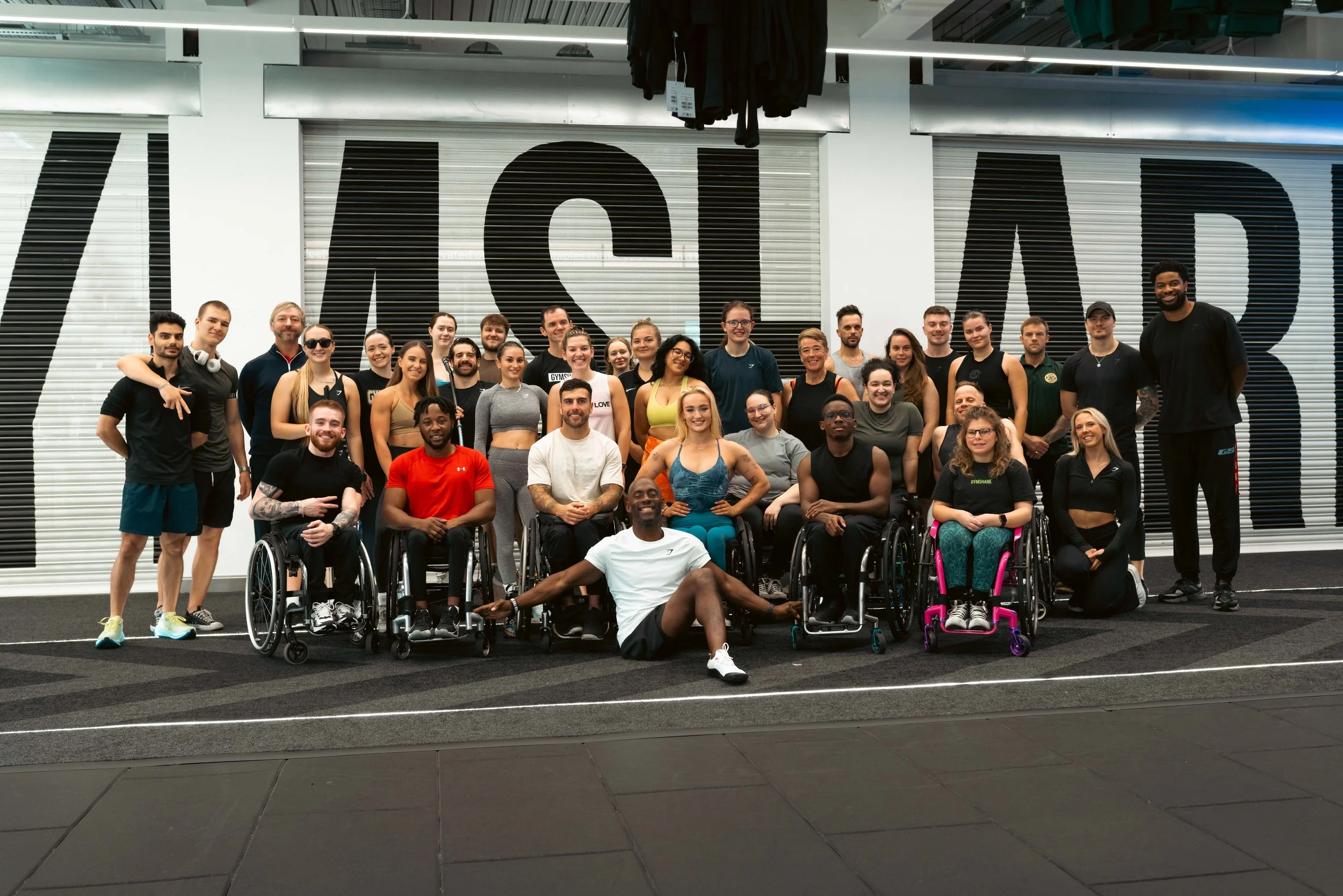 Group of diverse men and women, including individuals in wheelchairs, posed together in an indoor gym or fitness facility with a large sign in the background that says 'WASHO'.