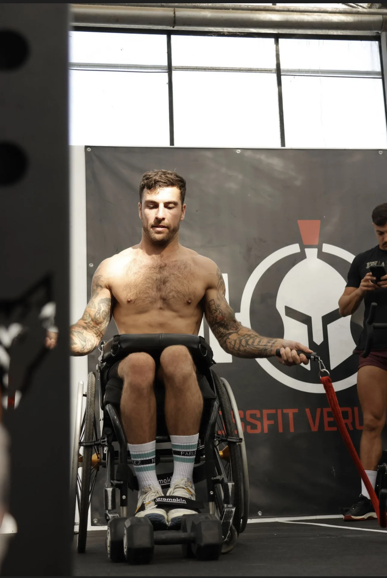 Male athlete with tattoos in a wheelchair during a CrossFit event, lifting weights, with a background banner displaying a CrossFit logo.