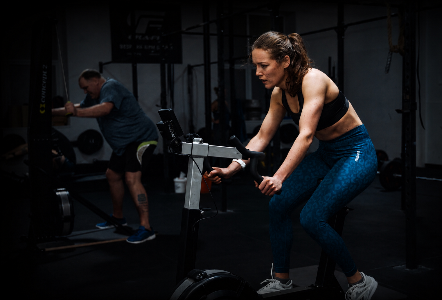 Member using a stationary bike during a fitness class at Pantheon Plymouth while another participant trains in the background, showcasing inclusive group training in a modern gym environment.