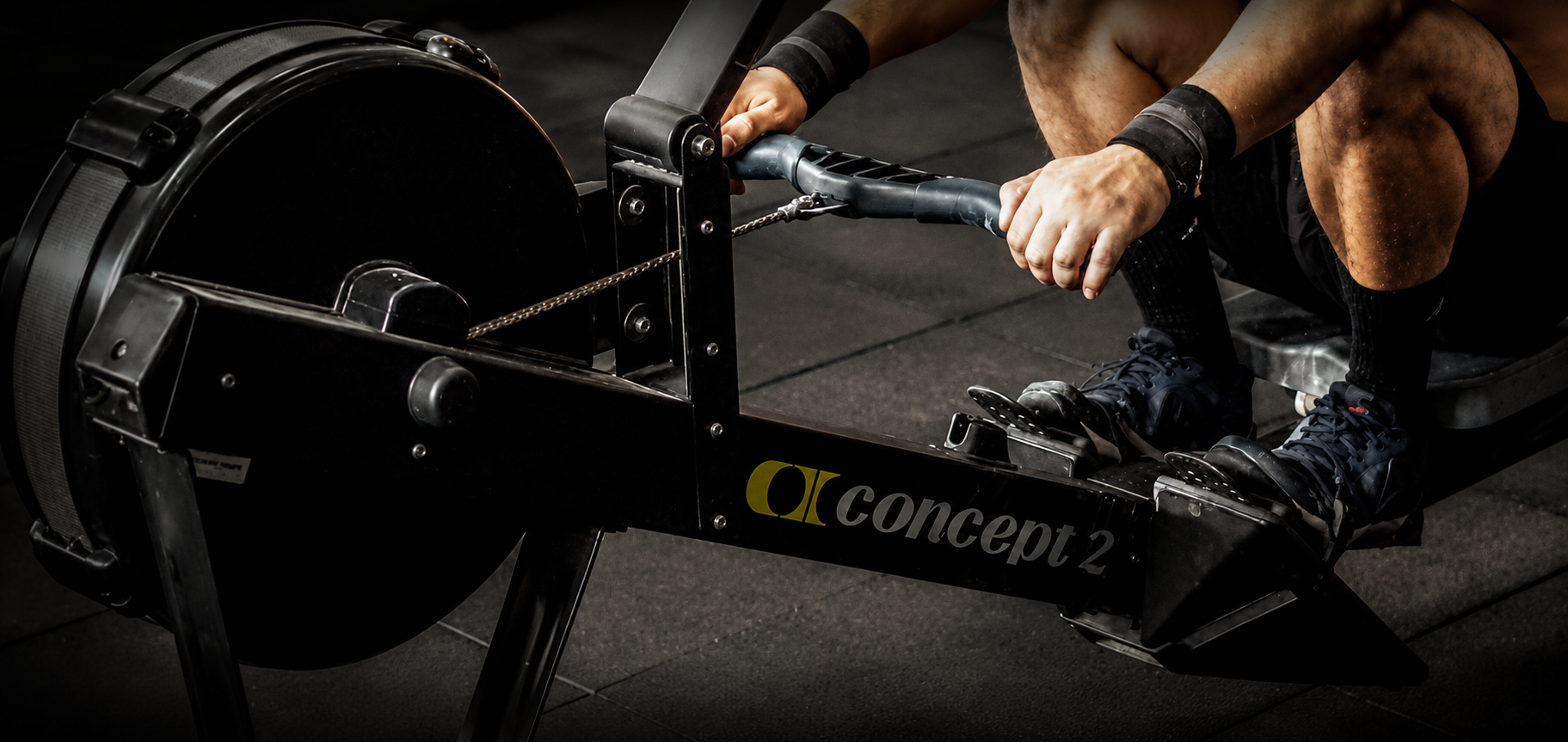 Person rowing a Concept2 indoor rowing machine, wearing sports shoes, black shorts, and wristbands, on a dark gym floor.