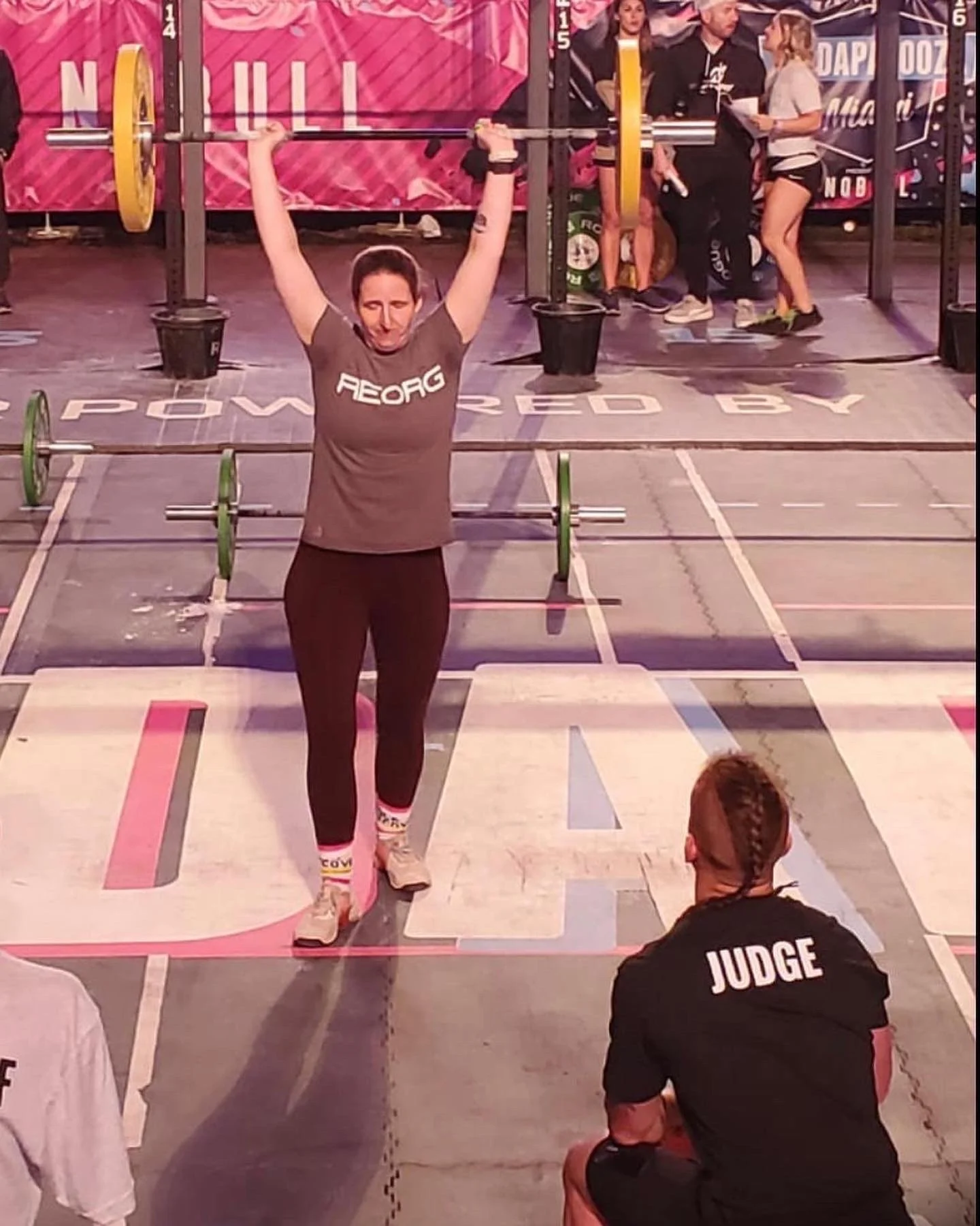 A woman in gray shirt lifting a barbell overhead during a weightlifting competition. She is wearing colorful socks. A judge in black with the word 'JUDGE' on the back is observing her. In the background, there are other people, some standing and talk
