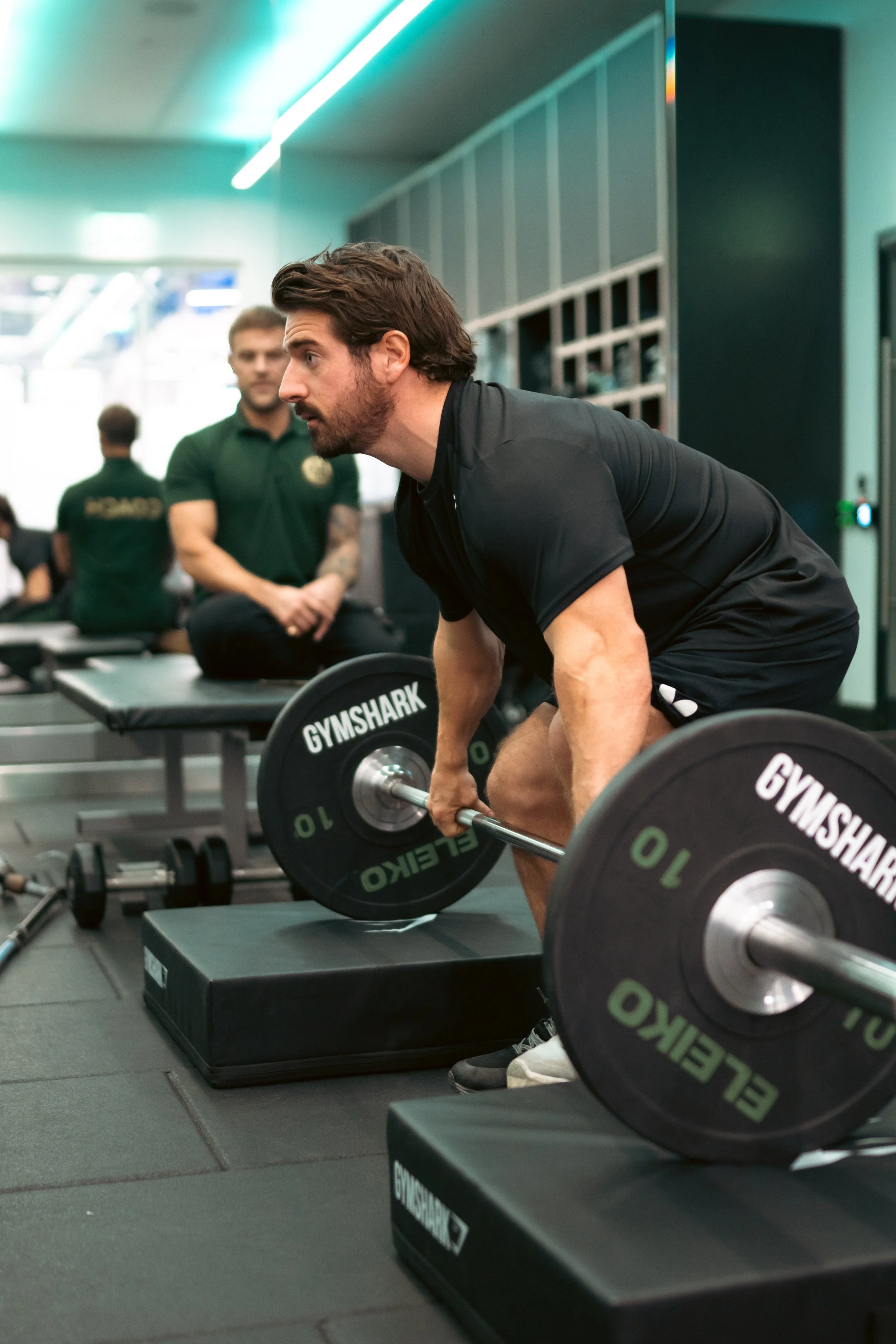 A man with dark hair and beard performing a deadlift exercise with a barbell in a gym, while others sit on benches in the background.