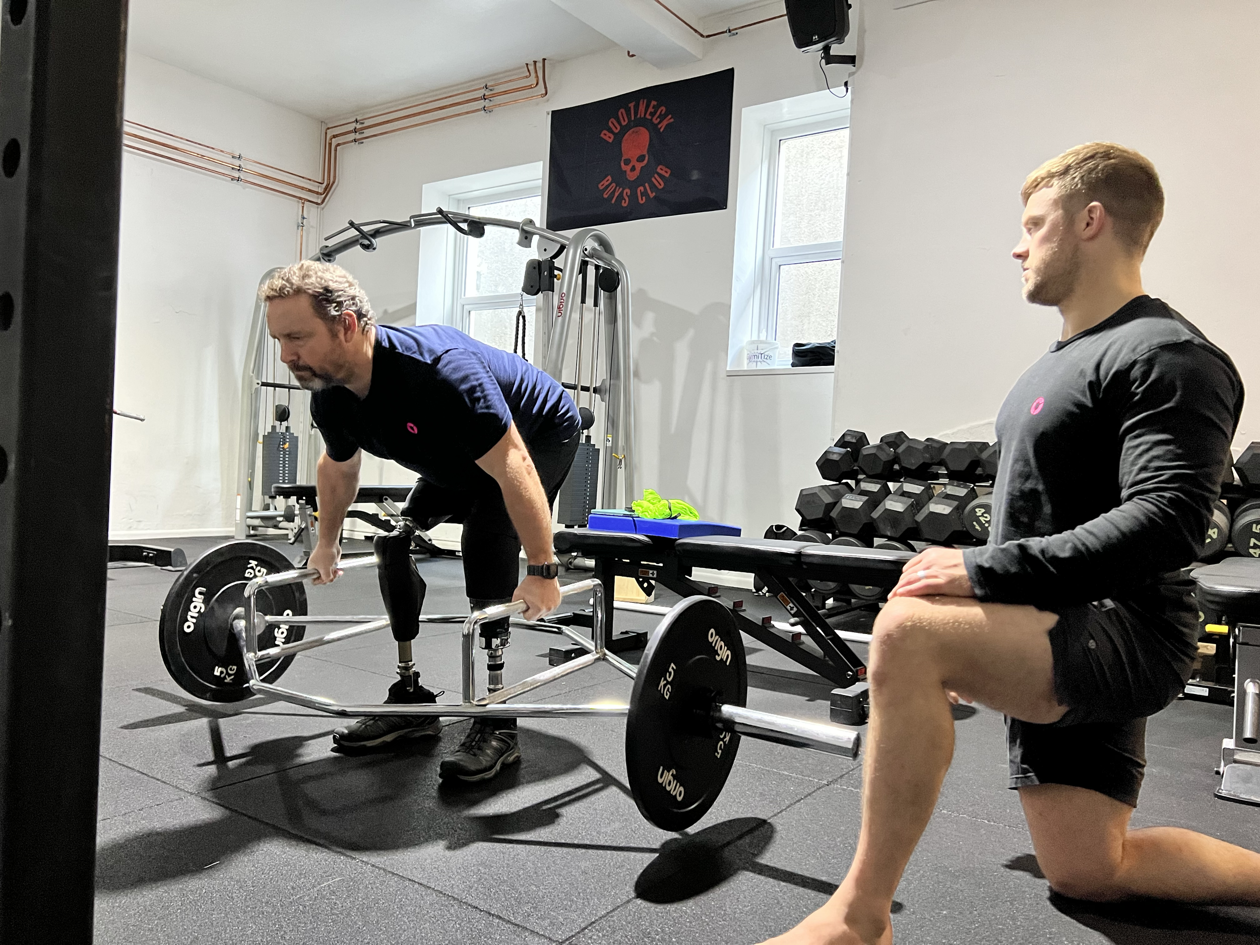 Two men working out in a gym. One man is doing an exercise with a barbell on a sled, while the other man is sitting on a bench observing. The gym has a black banner with a skull logo and the words "Bootsnek Boys Club" on the wall, along with other gy