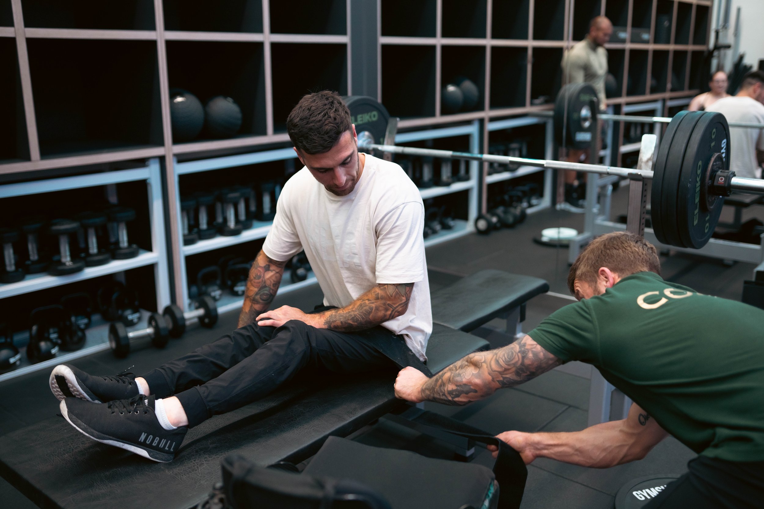 A man sitting on a treatment table receiving a leg massage from a therapist in a gym gym with weight storage and barbells in the background