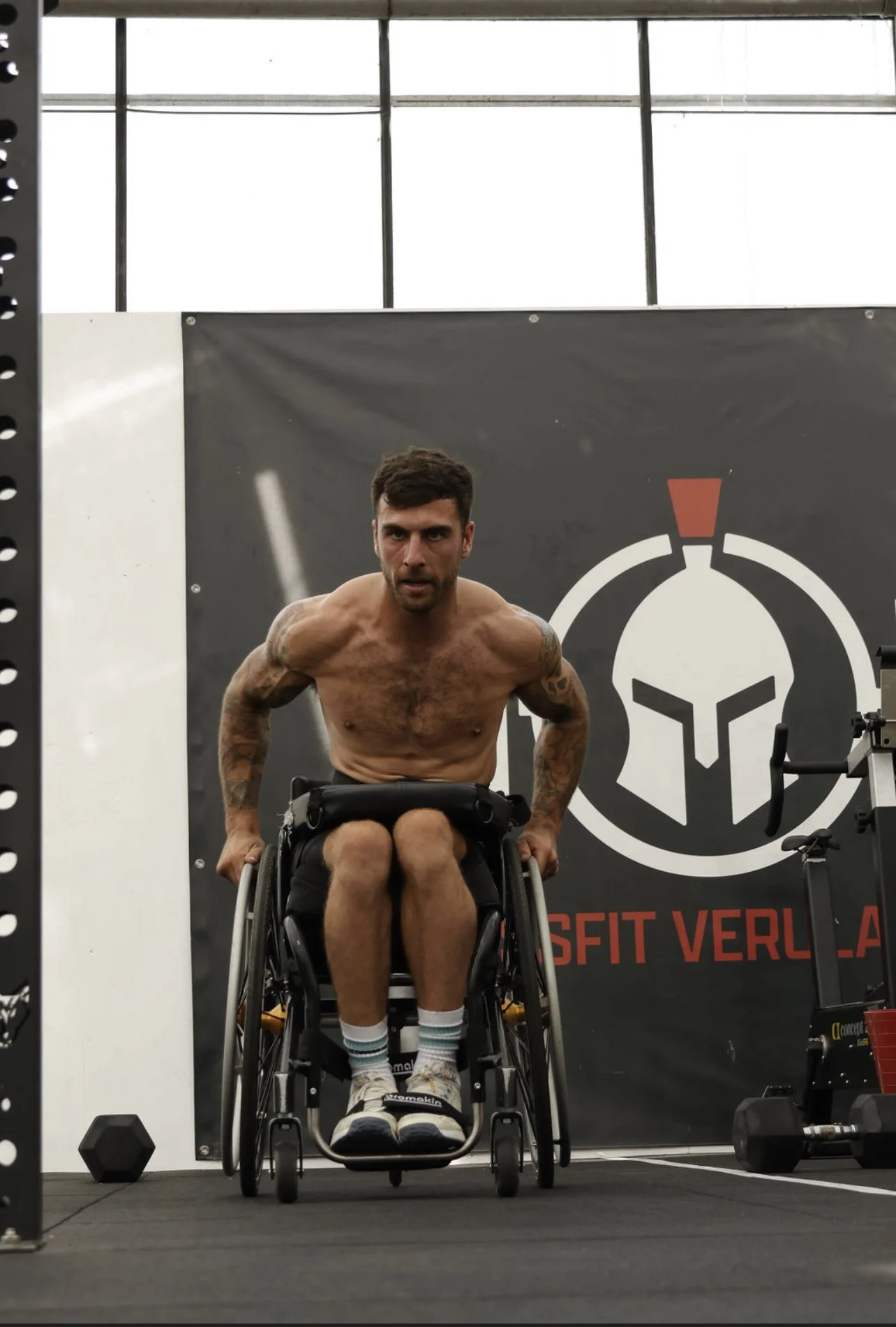 A shirtless man with tattoos in a wheelchair at a fitness gym, with a black logo on the wall behind him that says 'CrossFit Veracruz'.