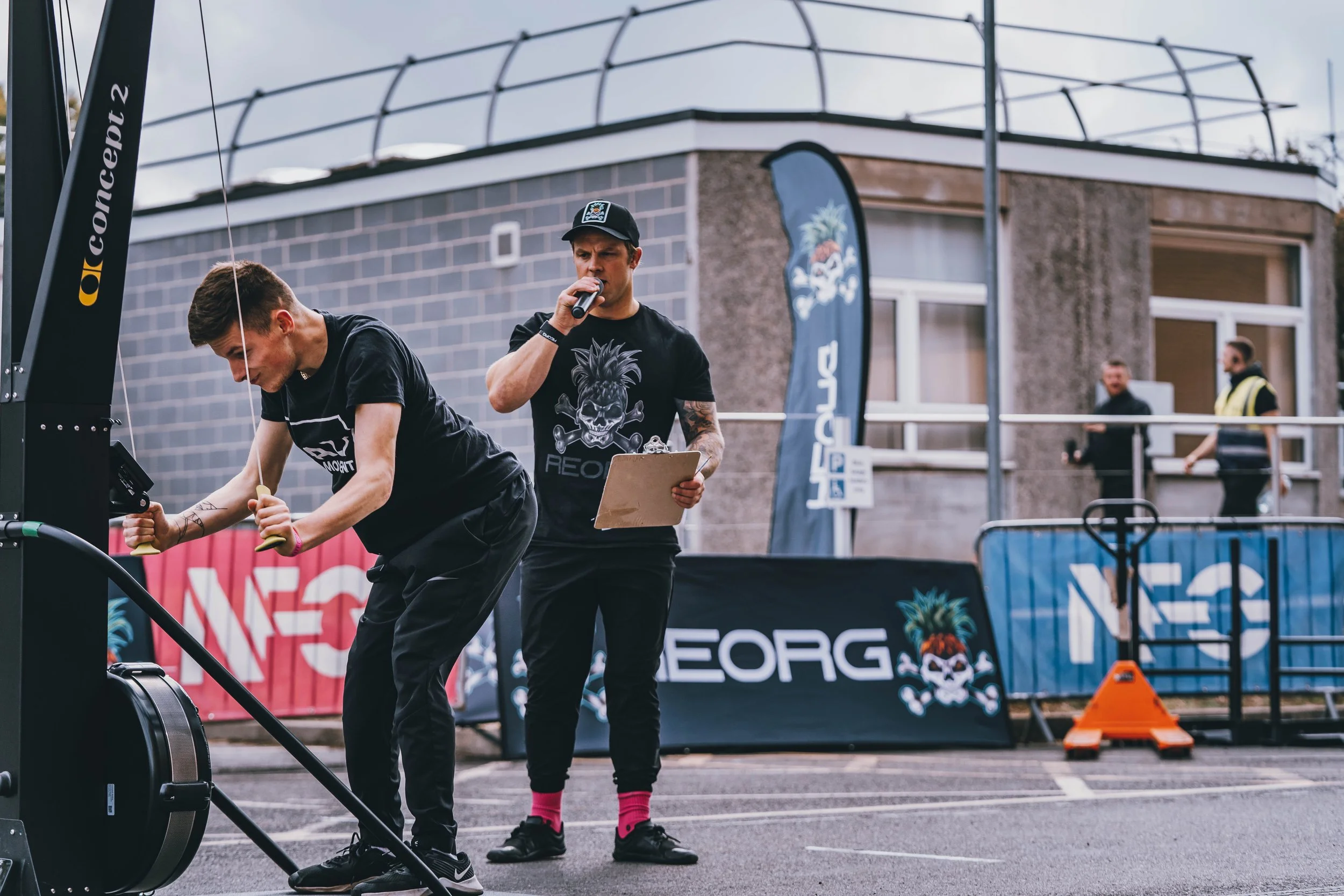 A man is using a rowing machine outdoors during a fitness event, with another man holding a microphone and clipboard nearby. In the background, there are banners, a building with large windows, and a few people watching.