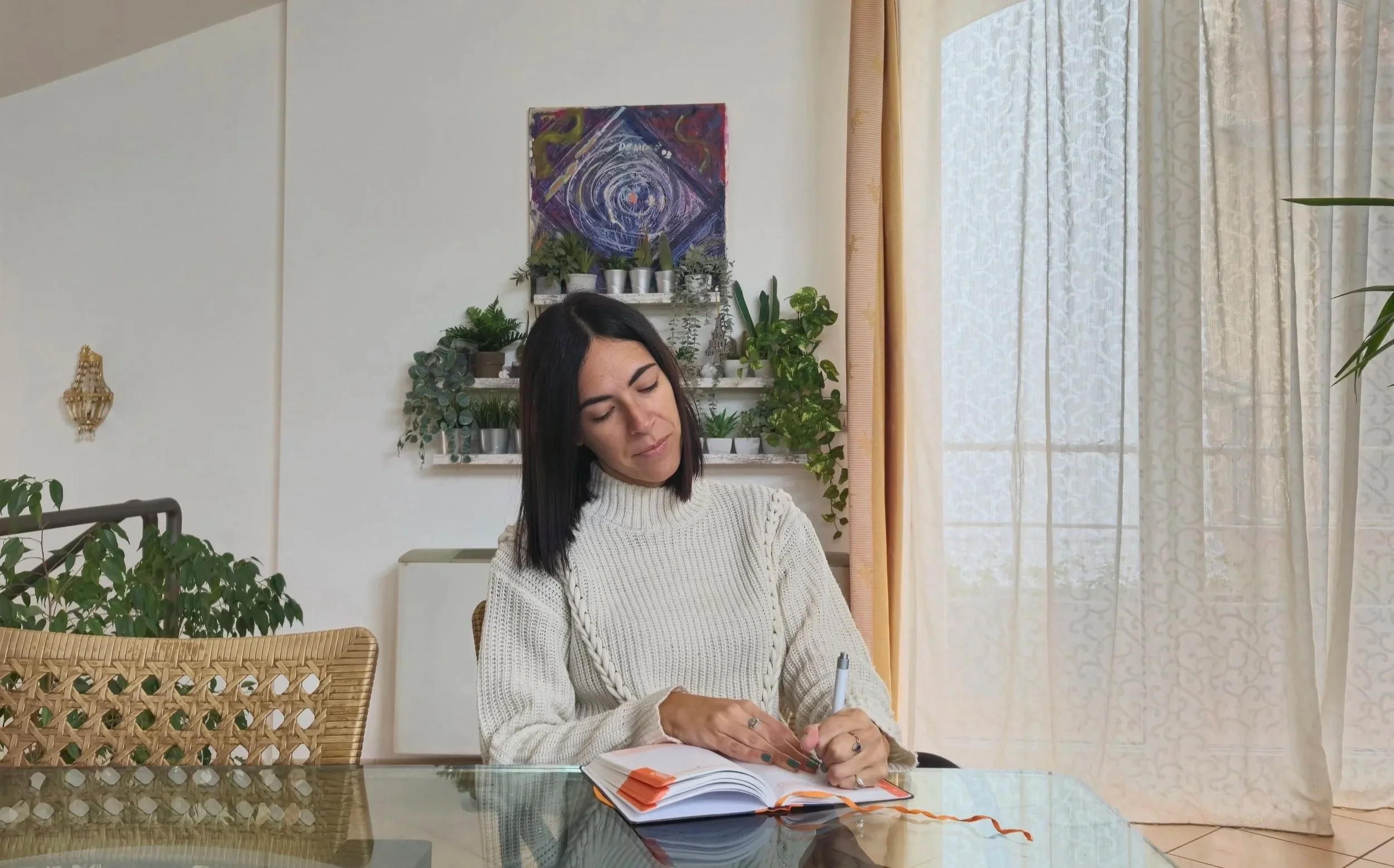 Una donna con capelli neri seduta a un tavolo di vetro, che scrive in un quaderno arancione, in un ambiente con molte piante e un quadro astratto sul muro