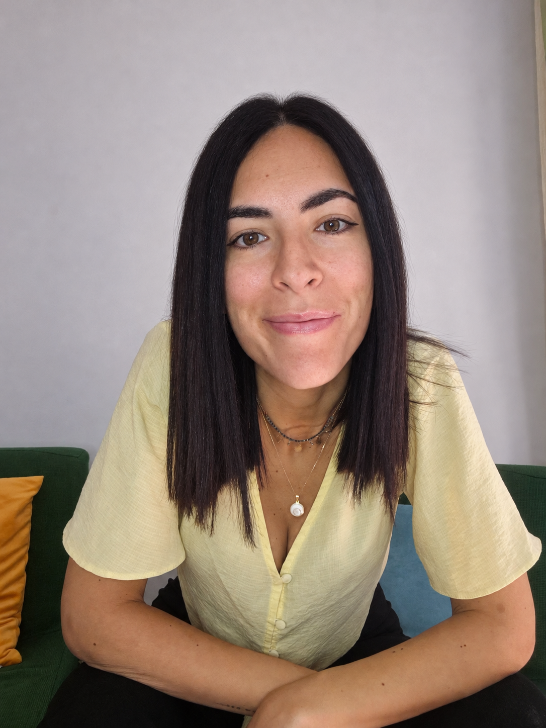 Una giovane donna con capelli neri, indossa una maglietta gialla e collane, sorride mentre si siede su un divano verde con cuscini arancioni e blu.