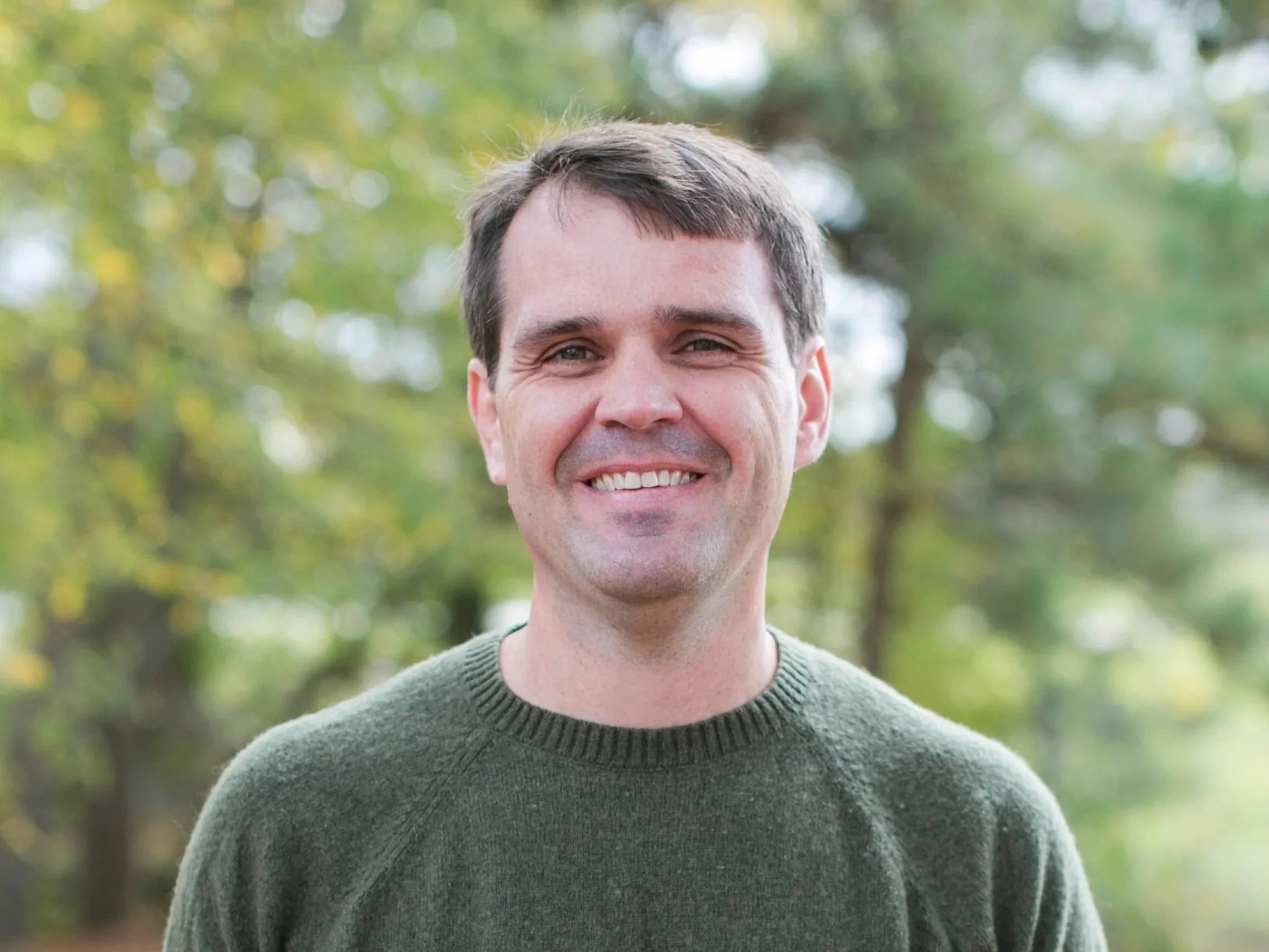 Erik Potter, a smiling man outdoors in a green sweater with a blurred background of trees and foliage.