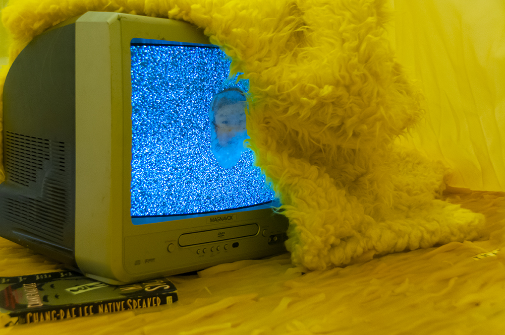  Is this | This is…  Multimedia installation: yellow curtains, old television, copy of Native Speaker by Chang Rae Lee, wig, 20 packs of rectangle dan moo ji, 5 packs of circle dan moo ji, print, shag rug, scrabble letters.  120” x 240” x 240”  Detai