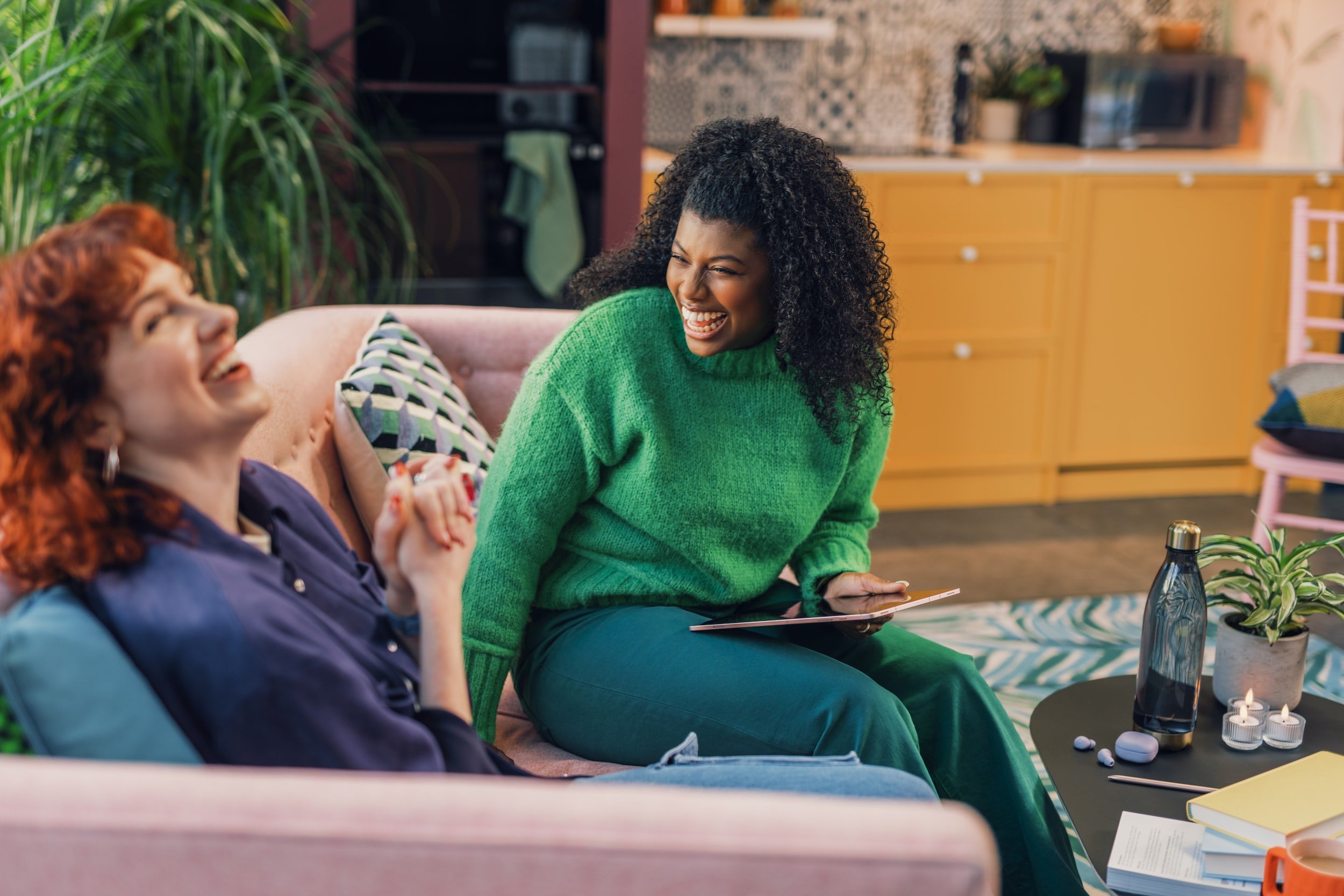 Two women laughing and chatting on a pink couch in a cozy living room. One woman is holding a tablet, and a small table in front has a plant, water bottle, and candles.