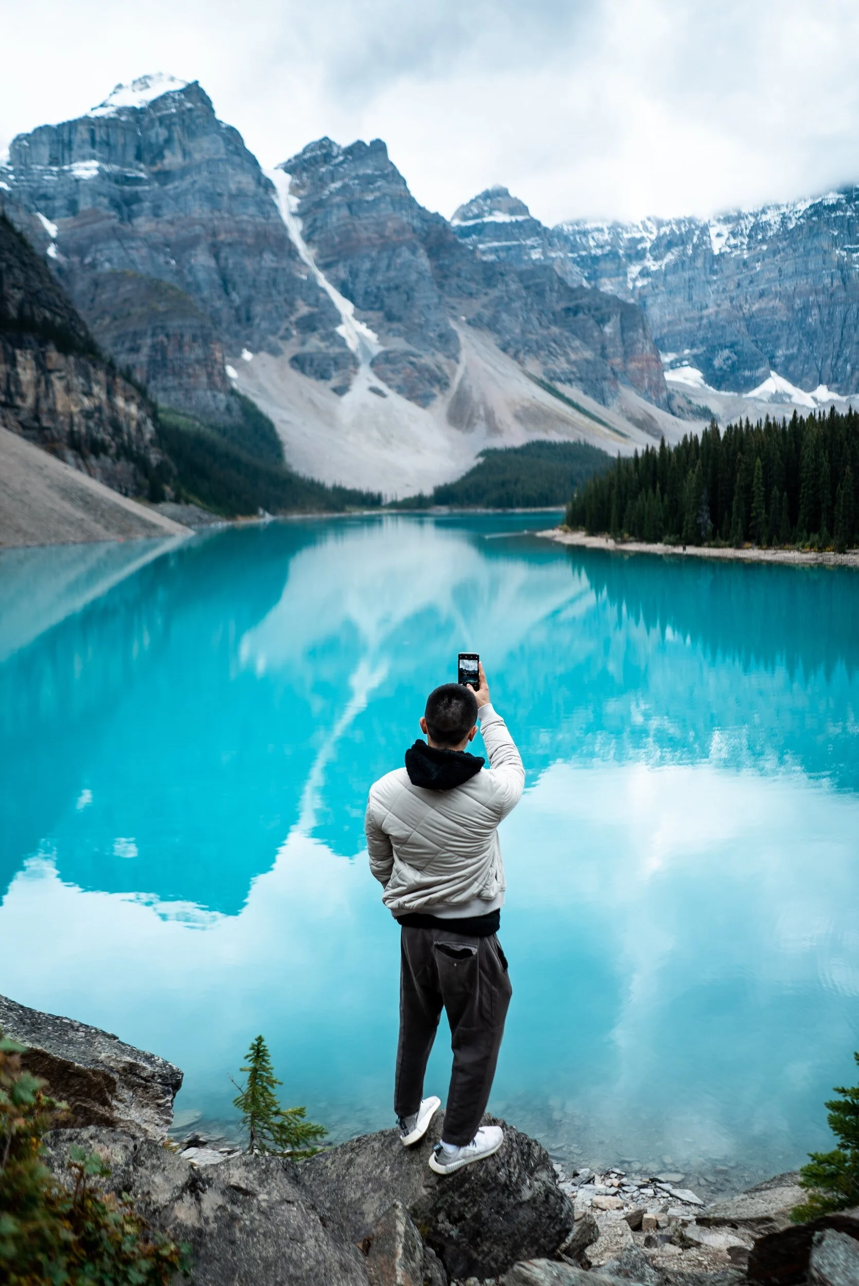 Man in a white jacket taking a photo of a turquoise lake with snow-capped mountains in the background.