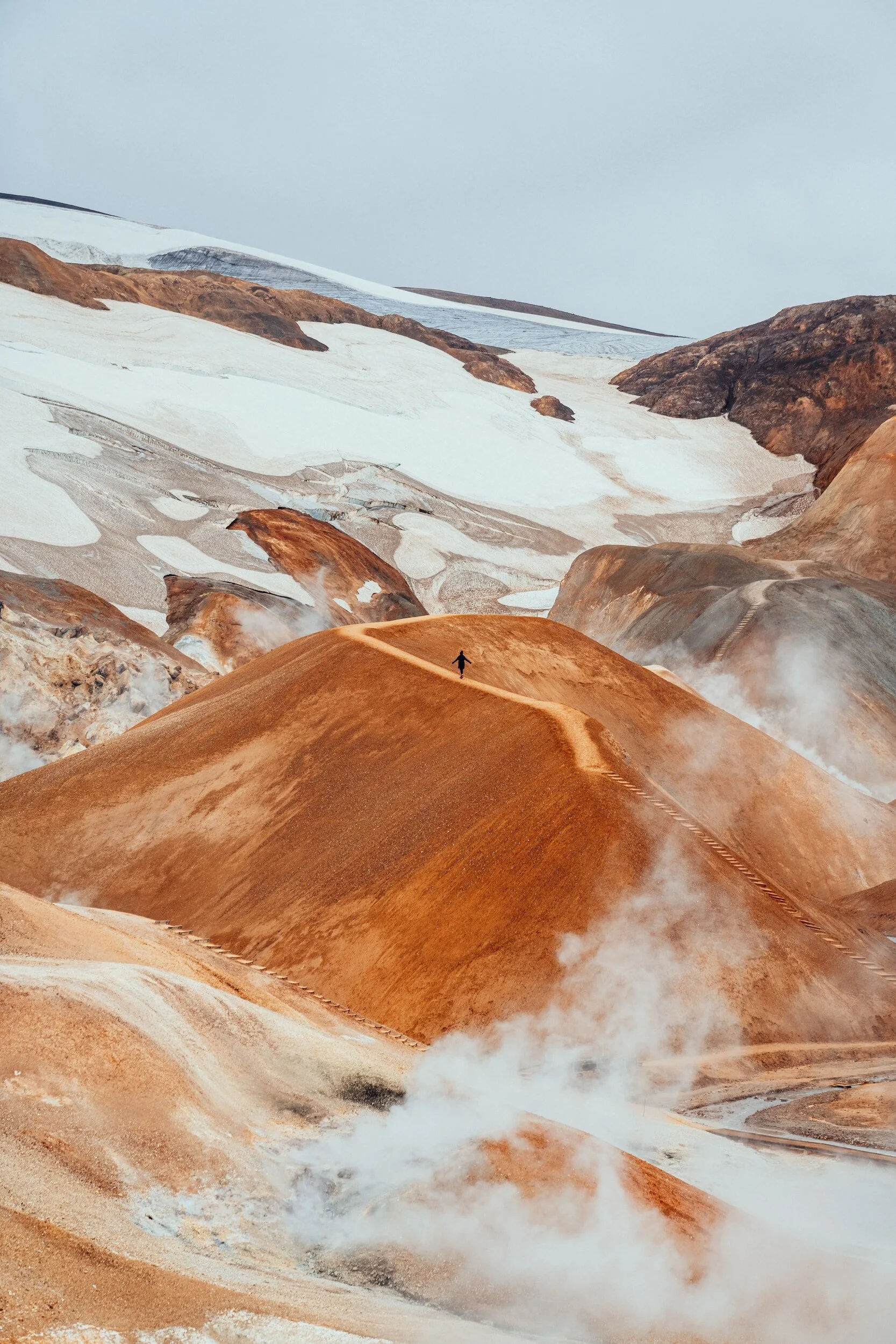 Person walking on a trail through a geothermal landscape with steaming vents, brown and orange hills, and patches of snow.