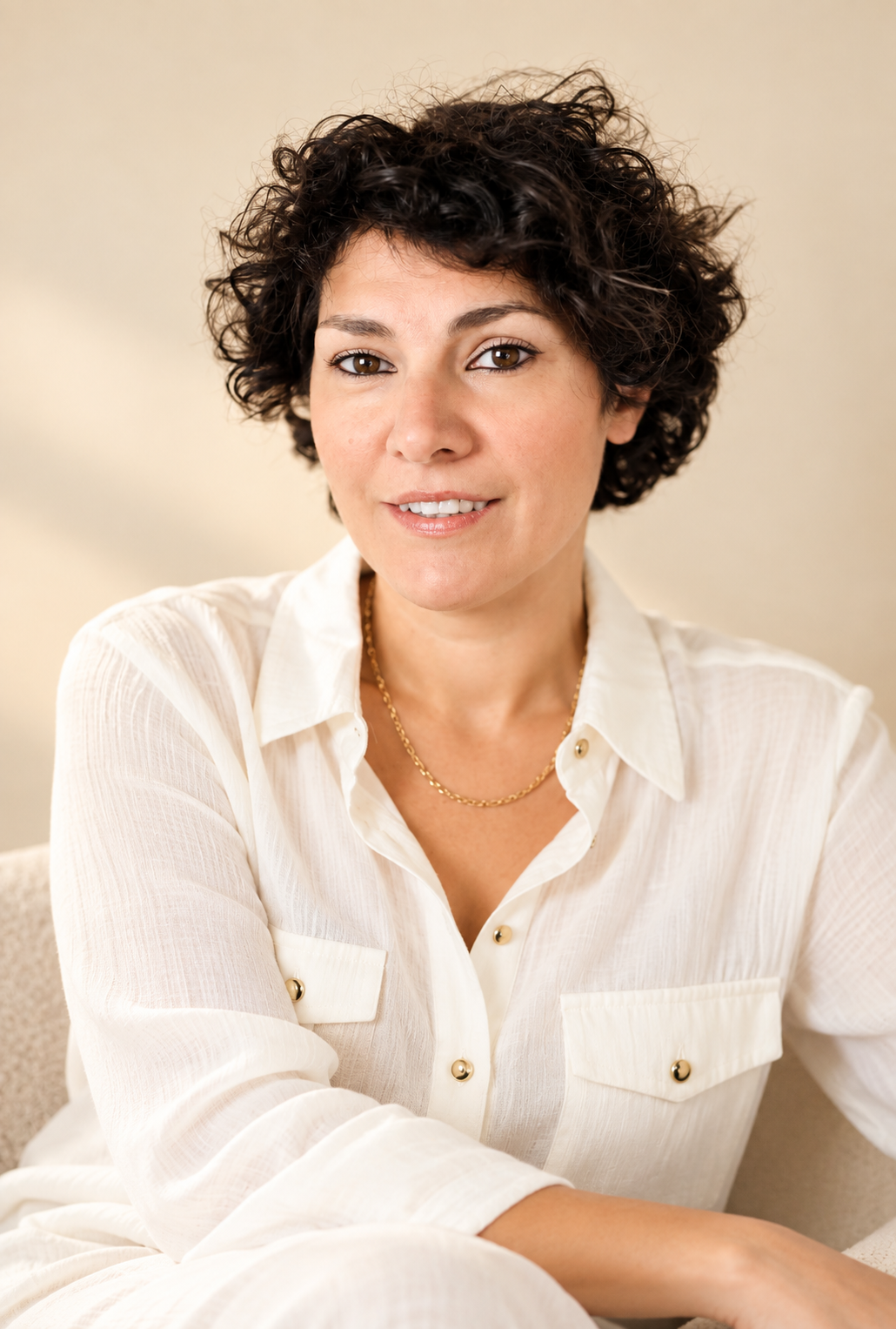 A woman with short, curly dark hair, wearing a cream-colored blouse with gold buttons and a gold chain necklace, sitting on a beige sofa, smiling softly at the camera.