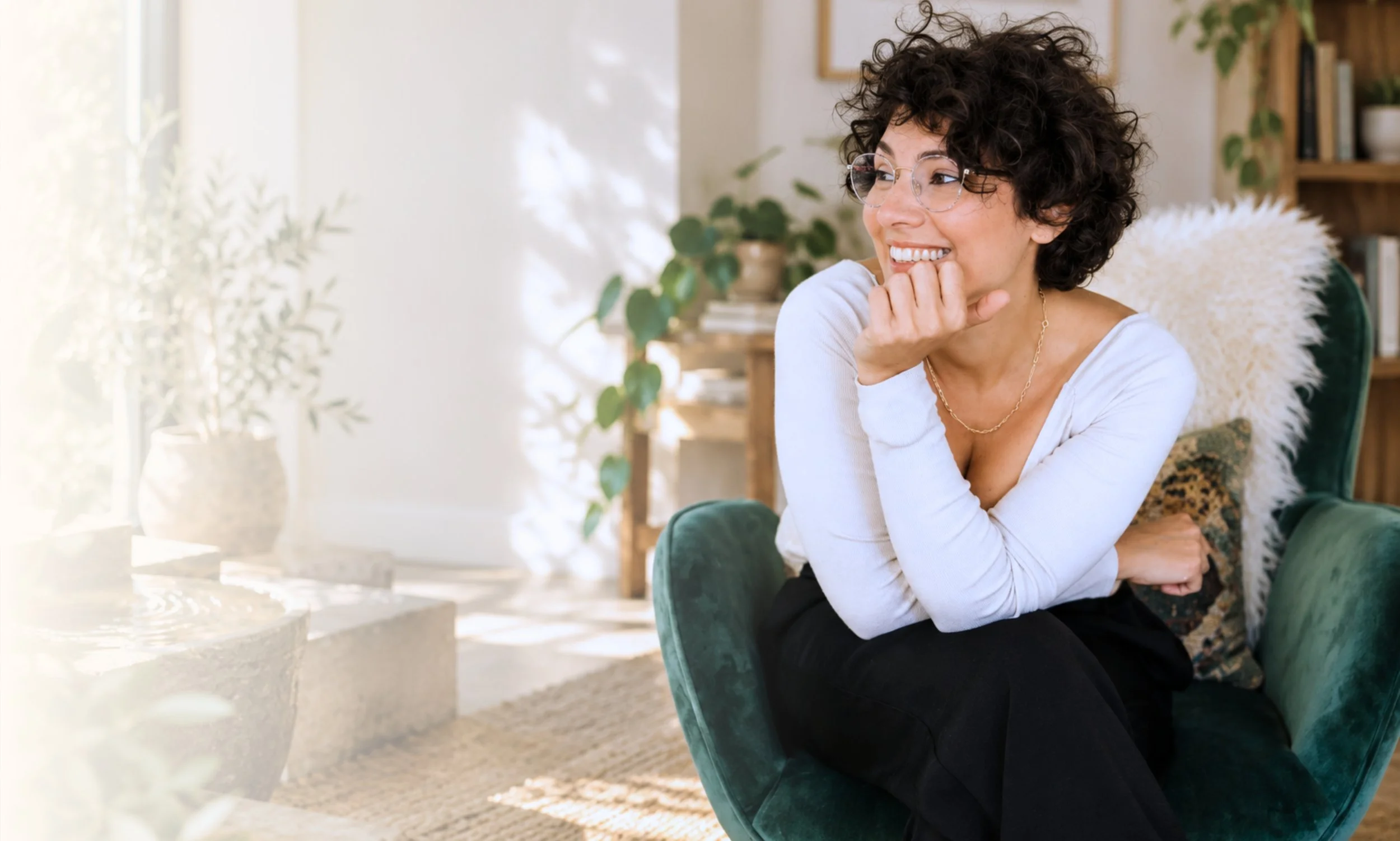 A woman with curly hair and glasses smiling while sitting in a green chair in a cozy living room with plants and bookshelves.