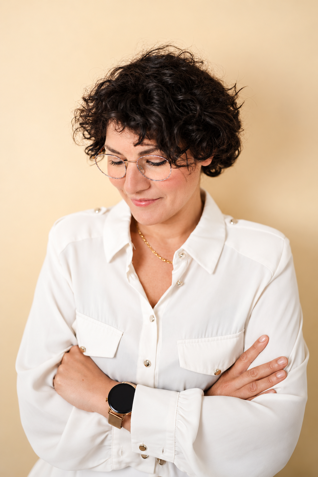 A woman with short, curly dark hair and glasses, wearing a white button-up shirt with rolled-up sleeves, standing against a light yellow background, with arms crossed and looking down.