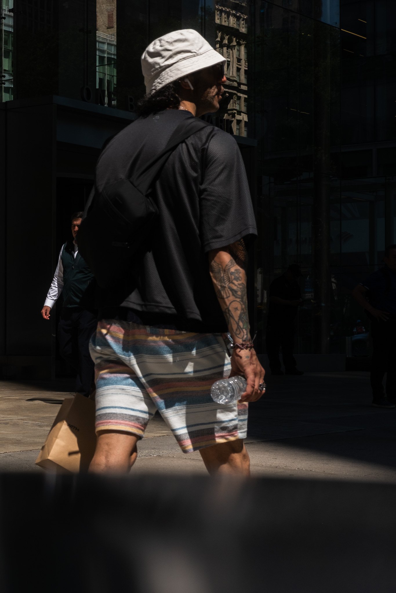 Street photograph of A person with tattoos, wearing a bucket hat, sunglasses, a black shirt, and striped shorts, holding a water bottle and a shopping bag, walking on a city street.