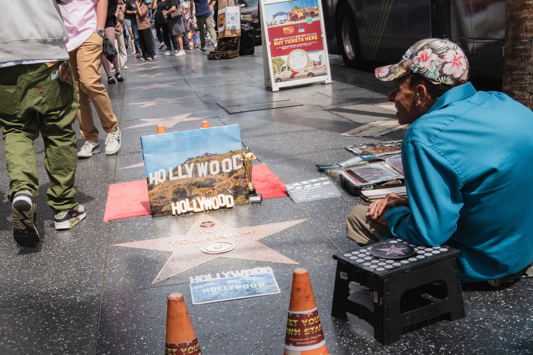 Street photograph of a street performer selling Hollywood souvenir photos and memorabilia on Hollywood Boulevard, California, with a star on the sidewalk and people walking by.
