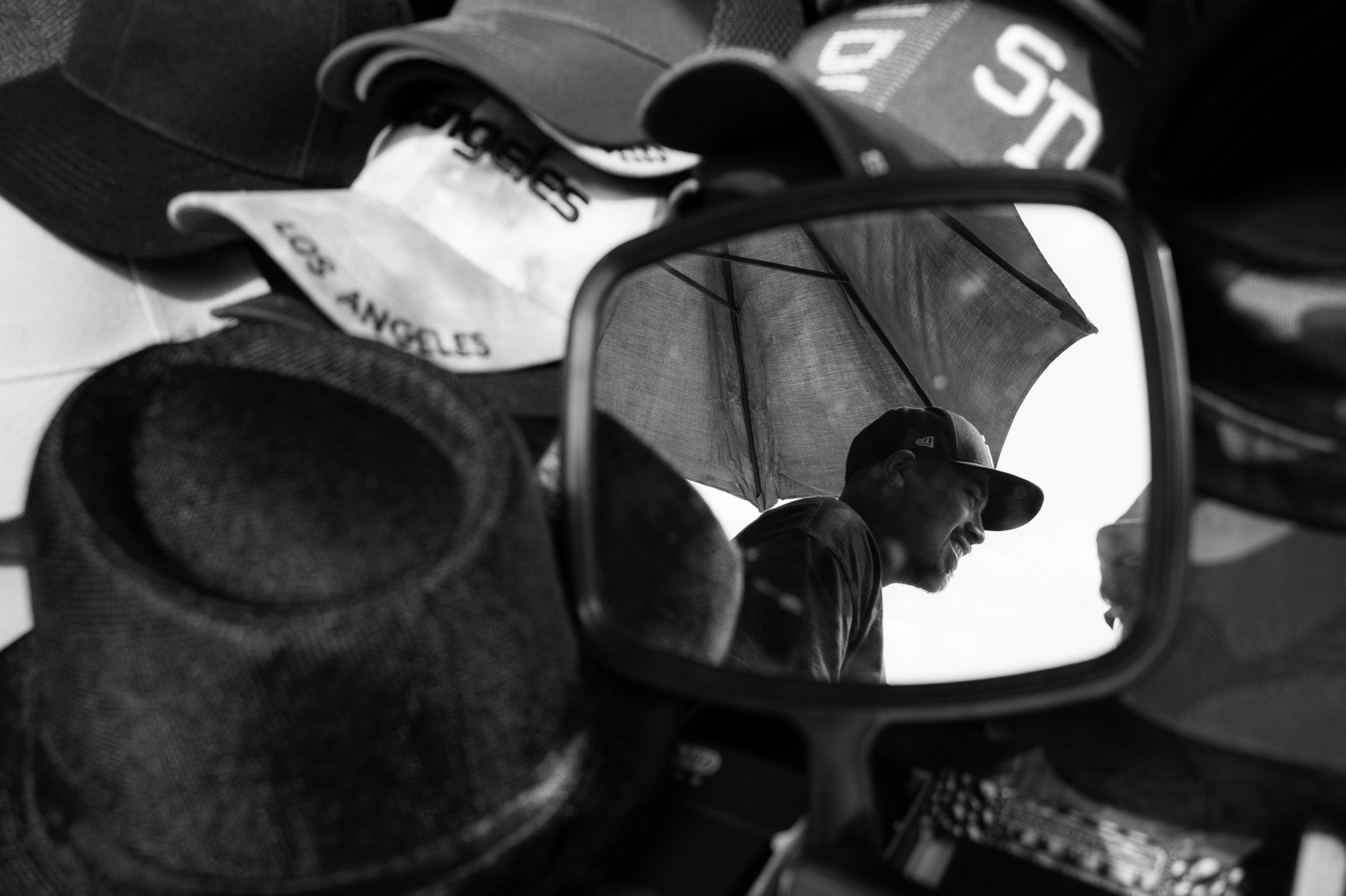 A man wearing a cap is seen smiling in a reflection of a mirror, with an umbrella above him. The mirror is surrounded by hats and clothing items.