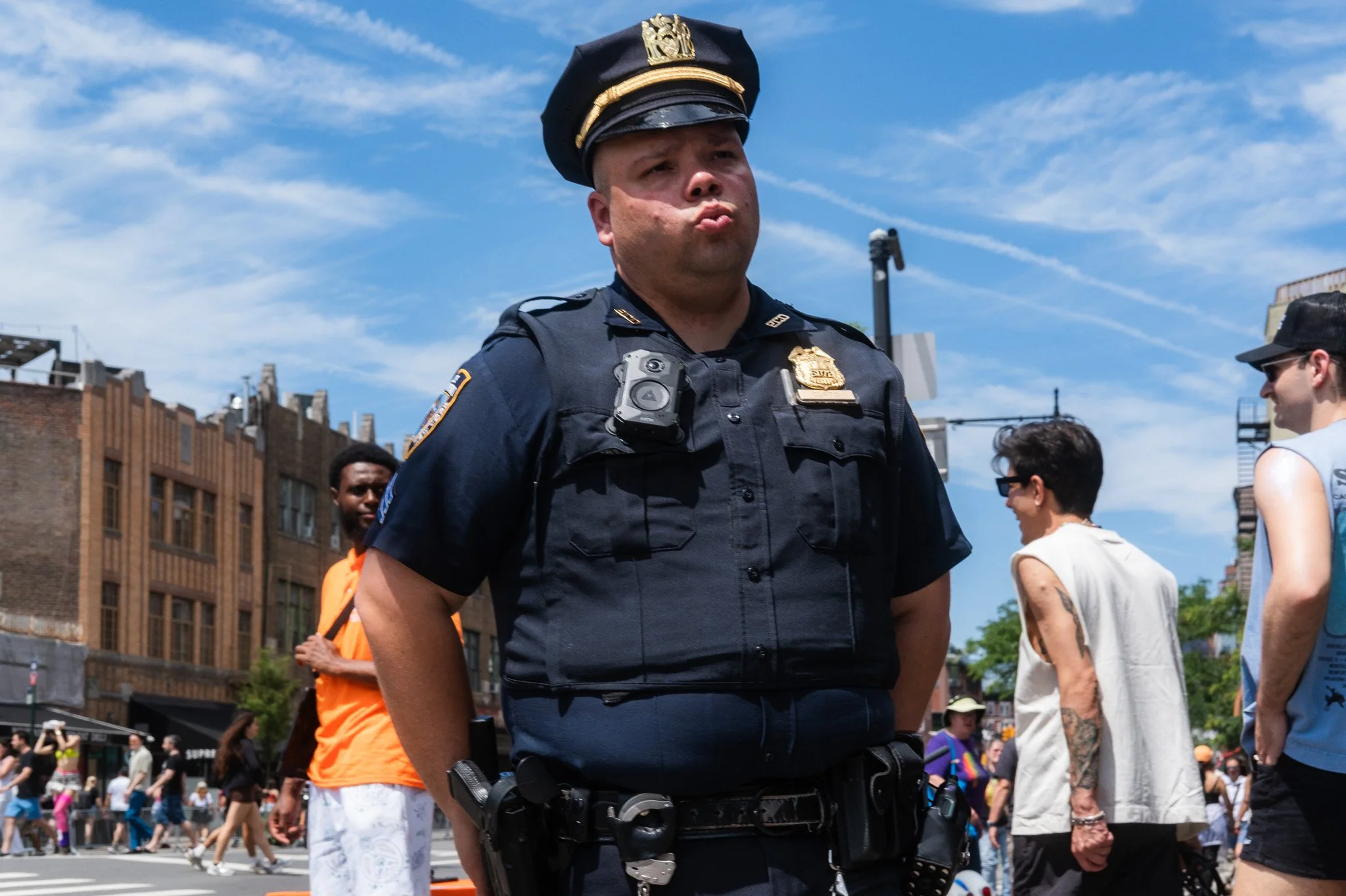 Street photograph of A police officer with a hat and sunglasses standing in a busy city street during a sunny day, with pedestrians behind him.