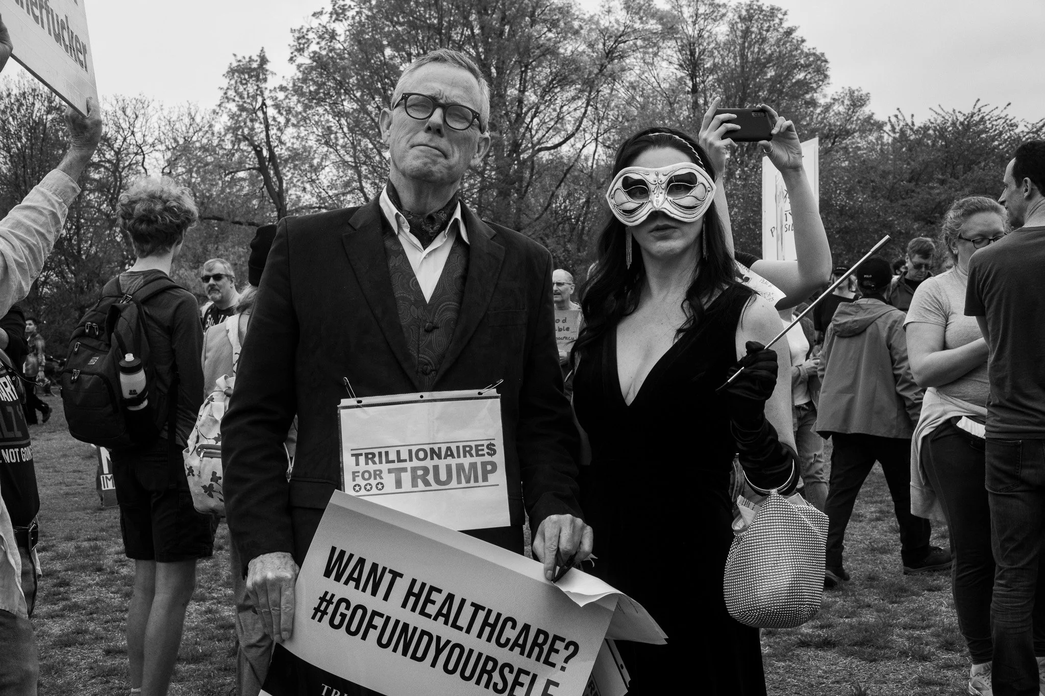 Two individuals at a protest rally, one holding a sign that reads 'Want Healthcare? #GOFINDYOURSELF' and the other wearing a mask and holding a small bag, with a crowd of people and trees in the background.